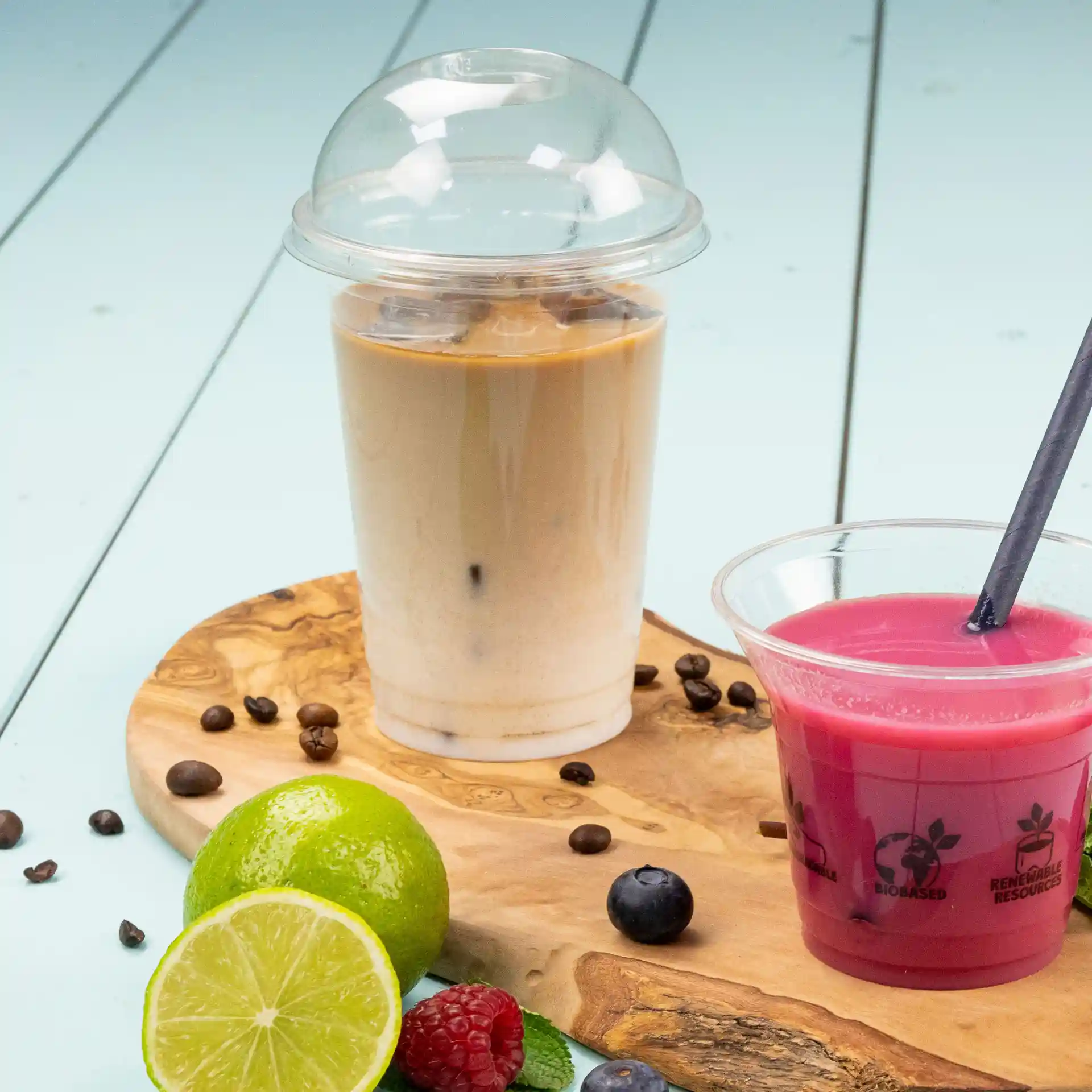 Afbeelding van een transparante 16 oz plastic beker gevuld met iced coffee, naast een roze drankje in een heldere beker op een houten plank met fruit.