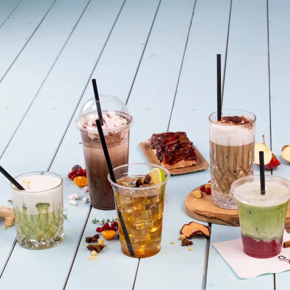 Verschillende dranken op een lichtblauwe houten oppervlakte; de dranken zijn een groene smoothie, een iced tea, een koffie en een dessert.