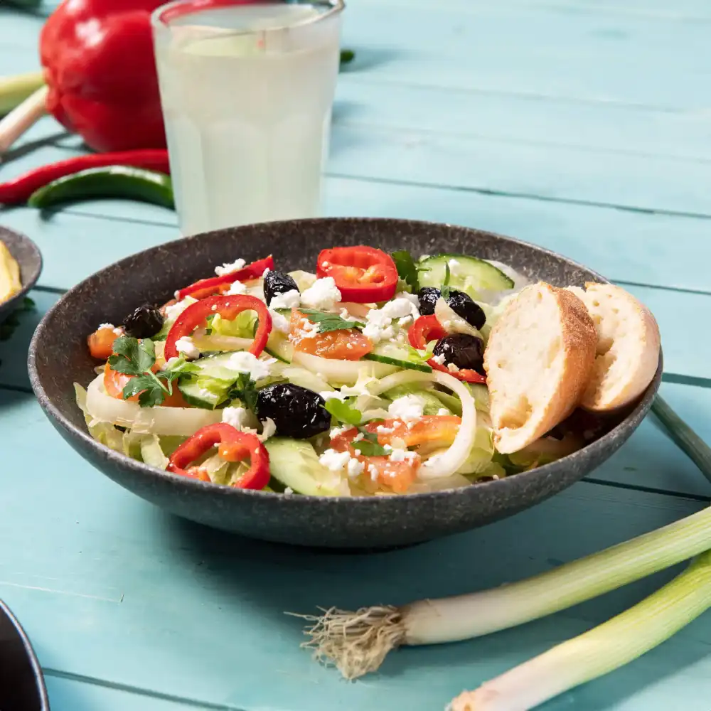 Kom met salade met komkommer, tomaten, olijven, feta en plakjes rode peper, vergezeld van twee stukken brood.