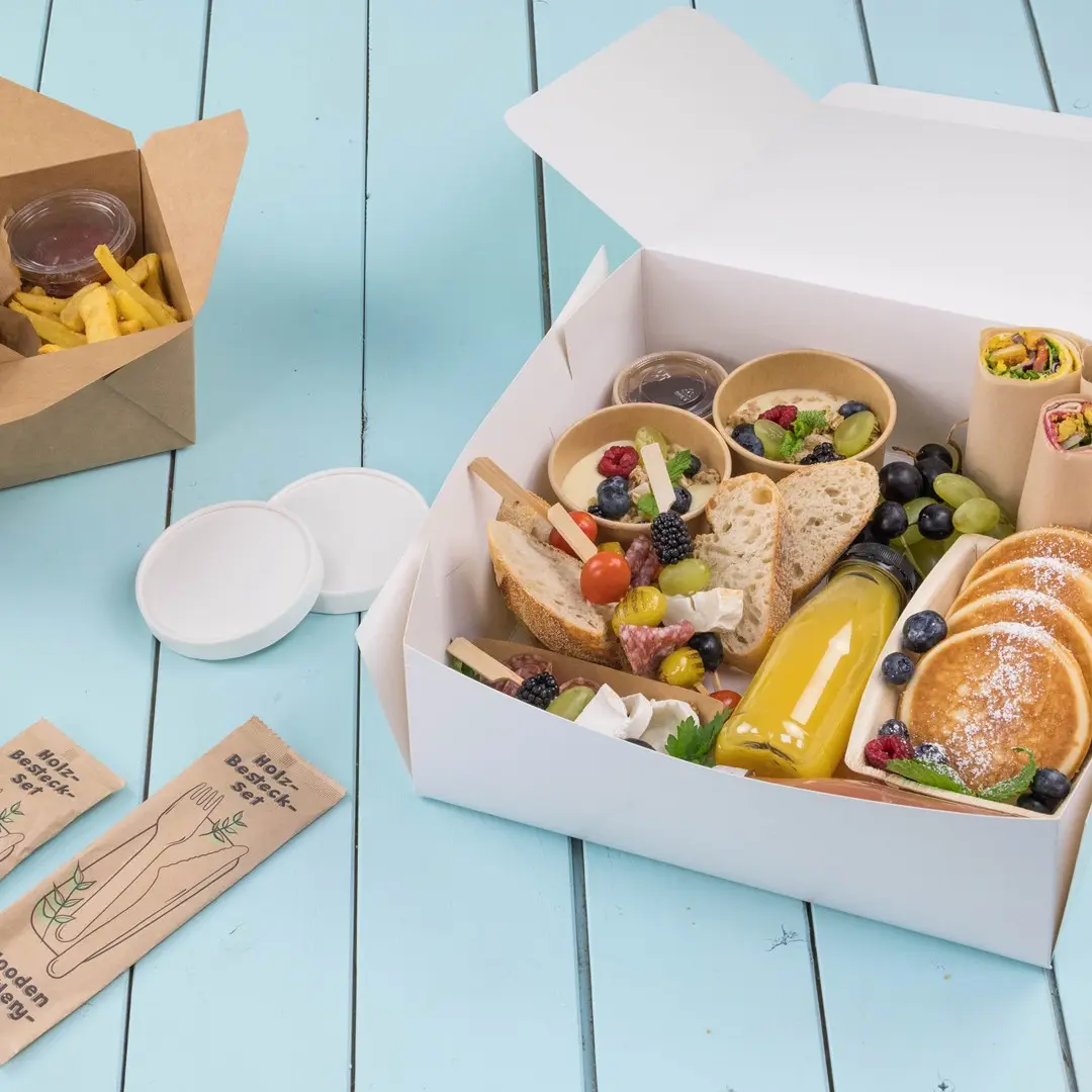 Witte take-away doos gevuld met verschillende voedingsmiddelen, waaronder brood, fruit, pannenkoeken en een drankje, op een blauwe houten ondergrond.