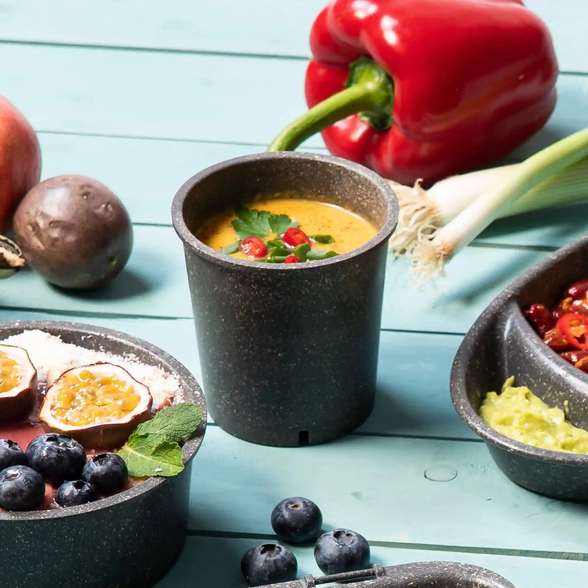 Ronde grijze herbruikbare beker met soep, gegarneerd met kruiden en chili, omringd door fruit en groenten op een blauwe ondergrond.
