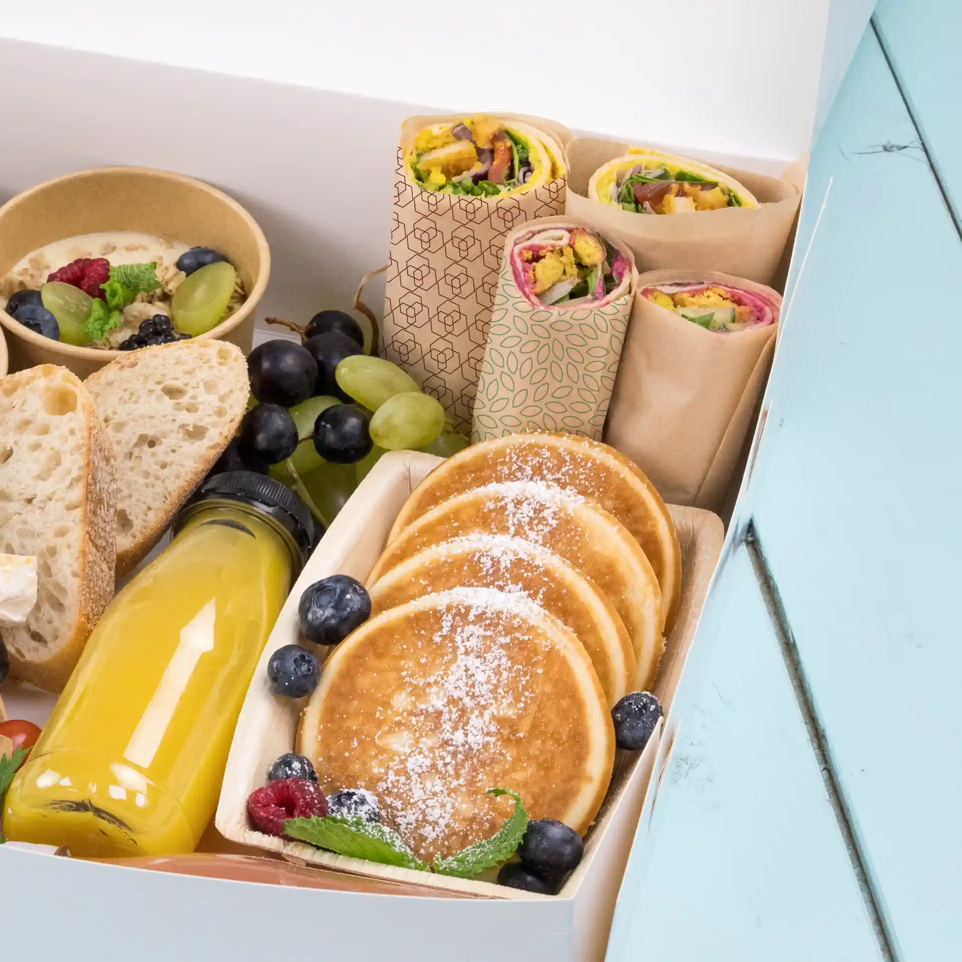 Diverse voedingsmiddelen zoals wraps, pannenkoeken, fruit en een drankje in een doos op een blauwe ondergrond.