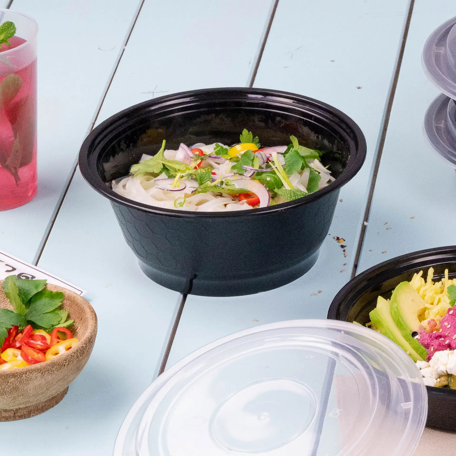 Een zwarte herbruikbare container gevuld met noedels, gegarneerd met verse kruiden en chili; andere voedingsmiddelen in de buurt.