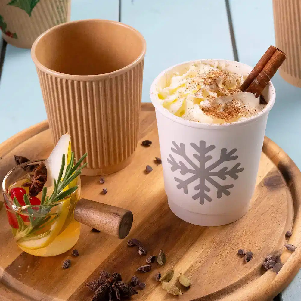 Afbeelding van een witte dubbelwandige papieren beker met een sneeuwvlokontwerp, gevuld met slagroom, naast een bruine geribde papieren beker en een versierde glas.