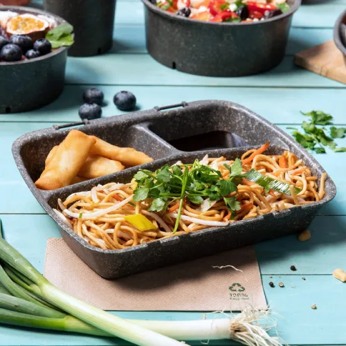 Maaltijdbox met noedels, groentegarnituur en loempia's, geplaatst op een blauwe houten tafel met verse ingrediënten eromheen.