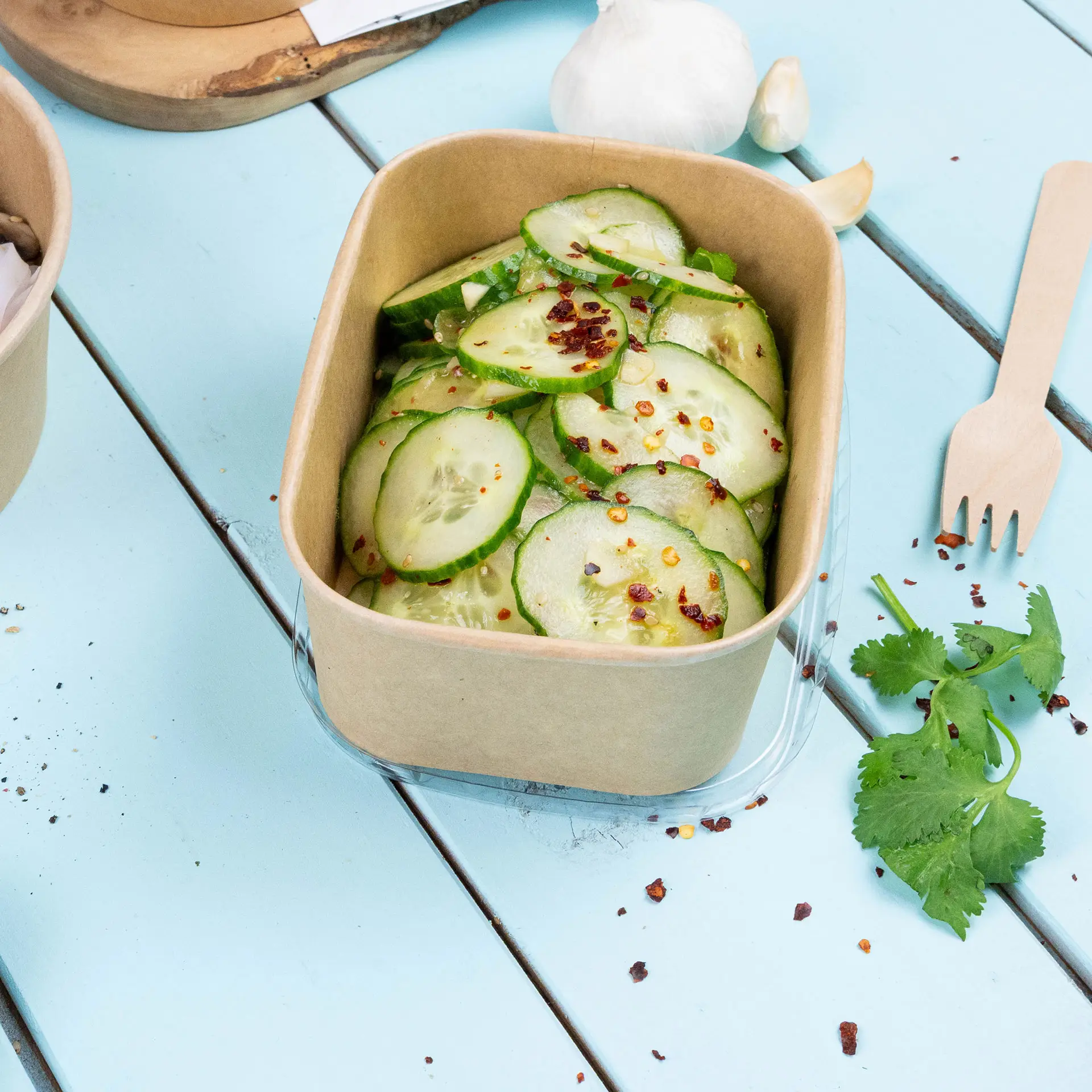 Bruine kraft voedselcontainer gevuld met gesneden komkommers, bestrooid met chilivlokken, op een lichtblauwe houten ondergrond.