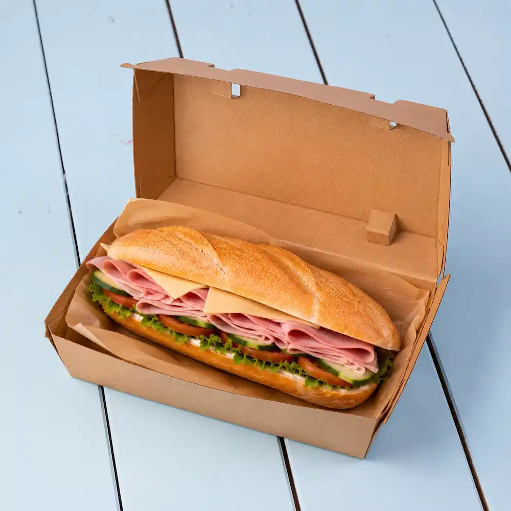 Sandwich in een bruine take-outdoos, met lagen ham, kaas, sla en gesneden komkommers.