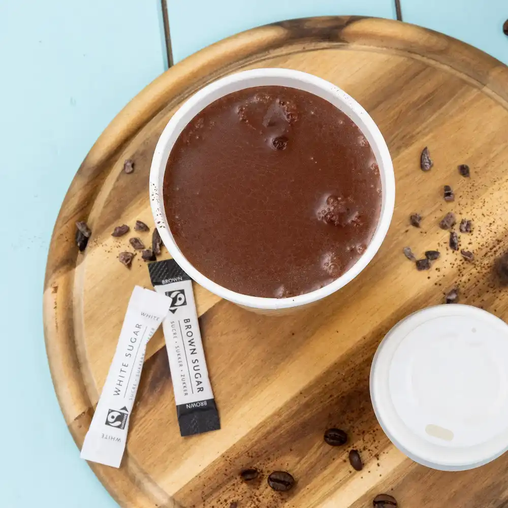 Warme chocolademelk in een witte beker met suikerzakjes, op een houten dienblad met koffiebonen en gemorste cacao.