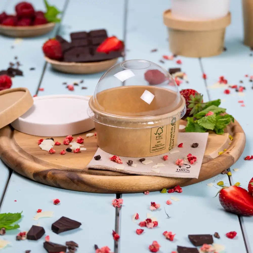 Afhaaltoetjescontainer met een transparant deksel op een houten dienblad, omringd door verse aardbeien, frambozen en chocoladestukjes.