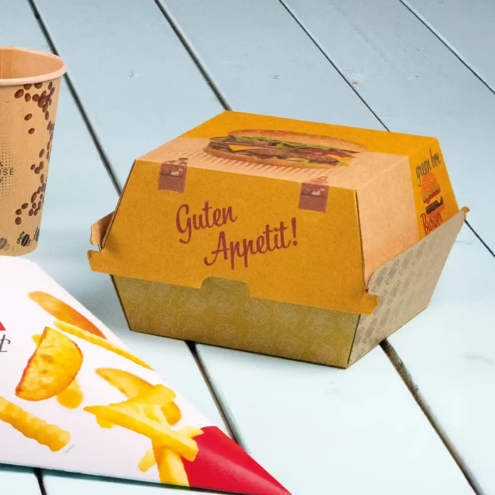 Bruine burgerdoos met de tekst "Guten Appetit!" naast een beker en een papier met fritesillustratie op een lichtblauwe ondergrond.