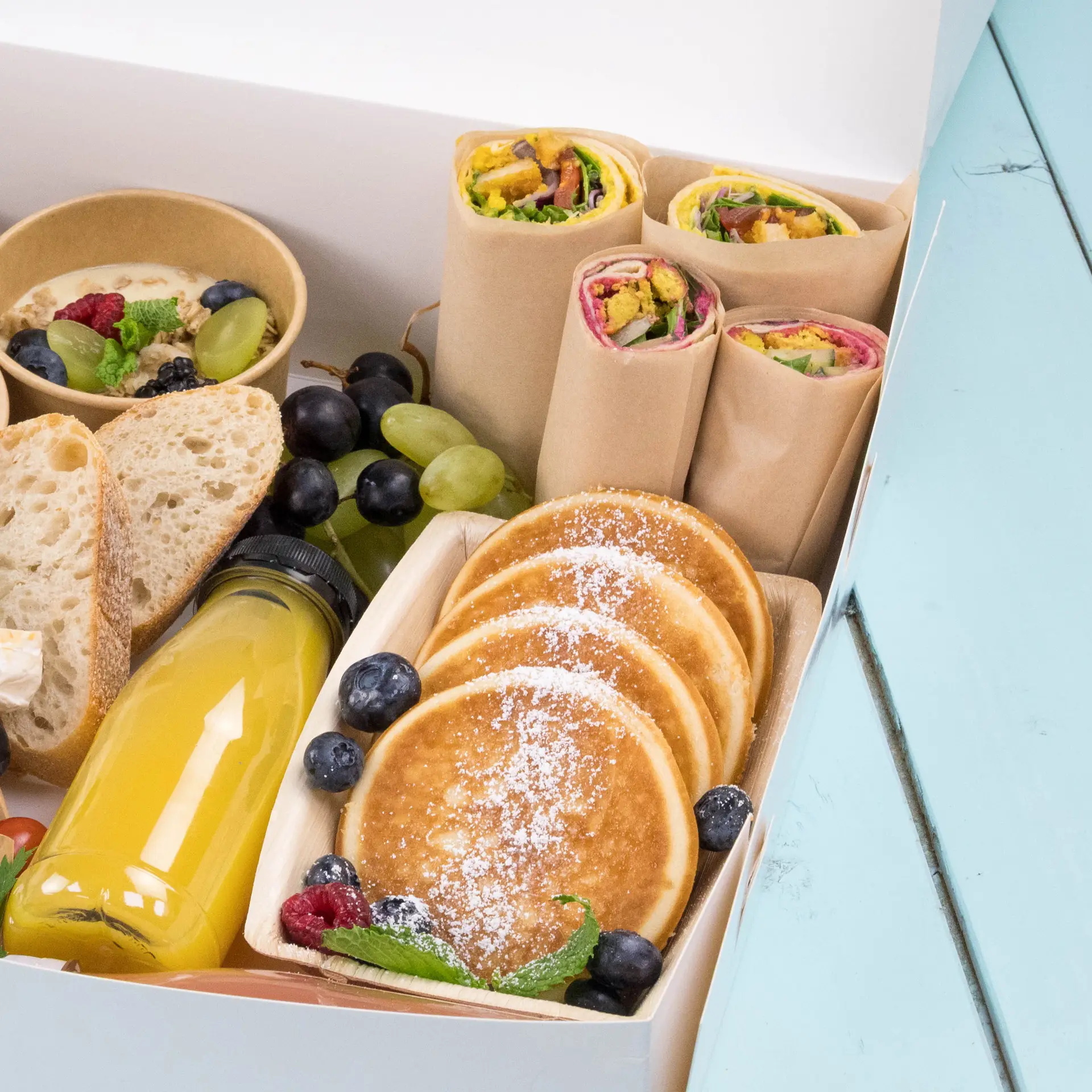 Diverse voedingsmiddelen waaronder pannenkoeken, wraps, brood, druiven en sap in een verpakkingsdoos.