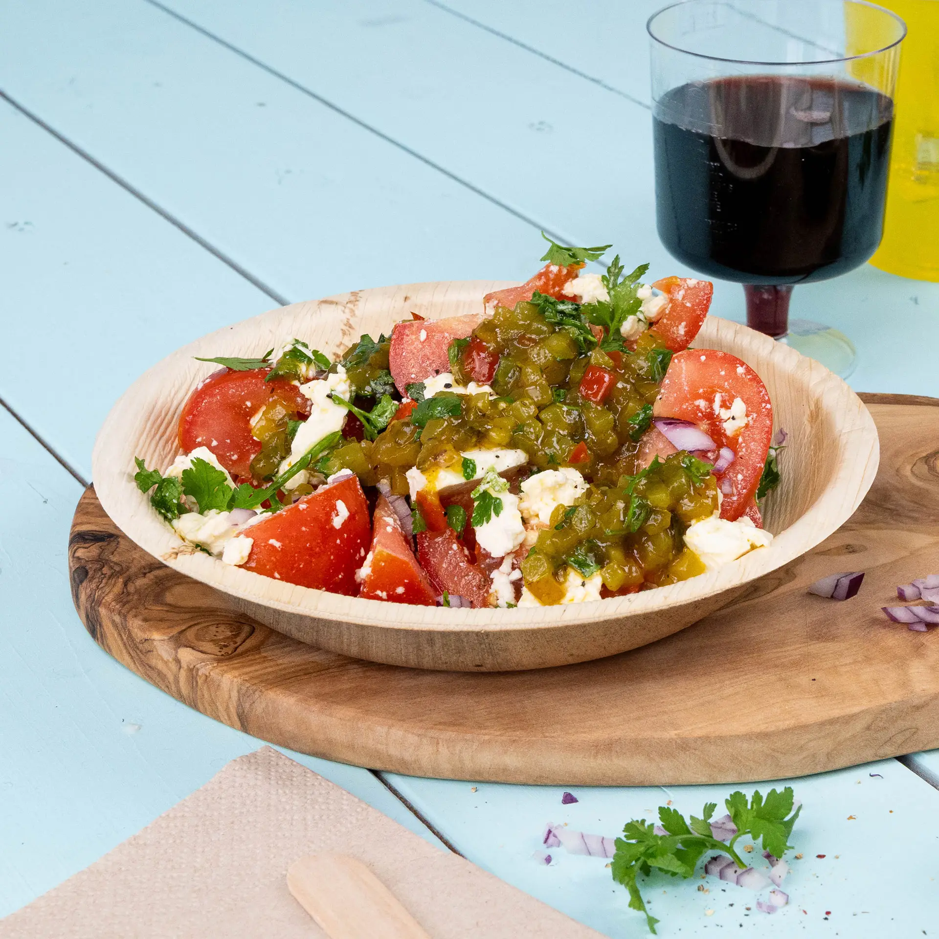 Een diep, rond palmblad bord gevuld met een salade van tomaten, kruiden en jalapeño, gezet op een houten plank met een drankje.
