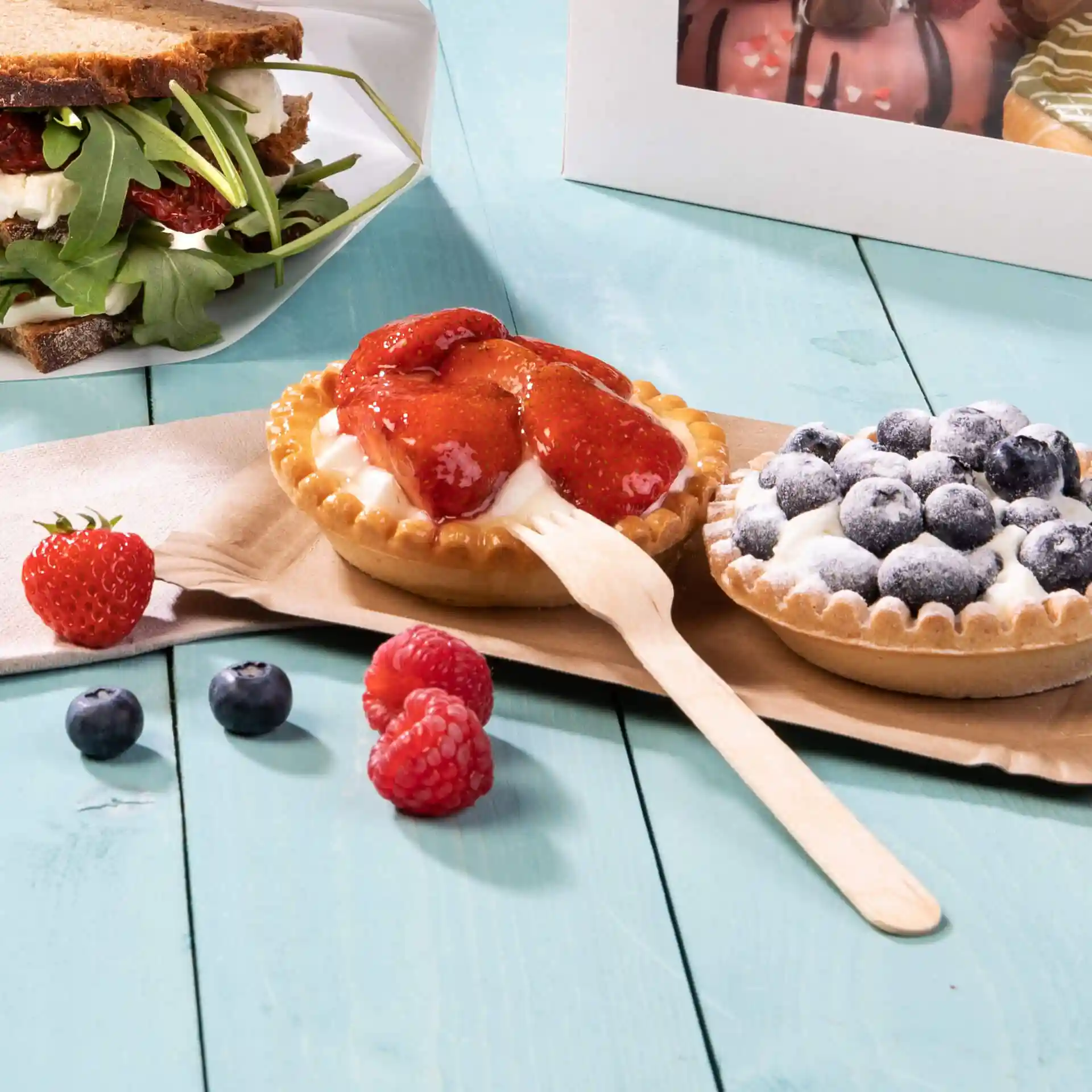 Afbeelding van twee desserttaarten met aardbei- en bosbessentopping, vergezeld van verse bessen en een houten vork op een lichtblauwe ondergrond.