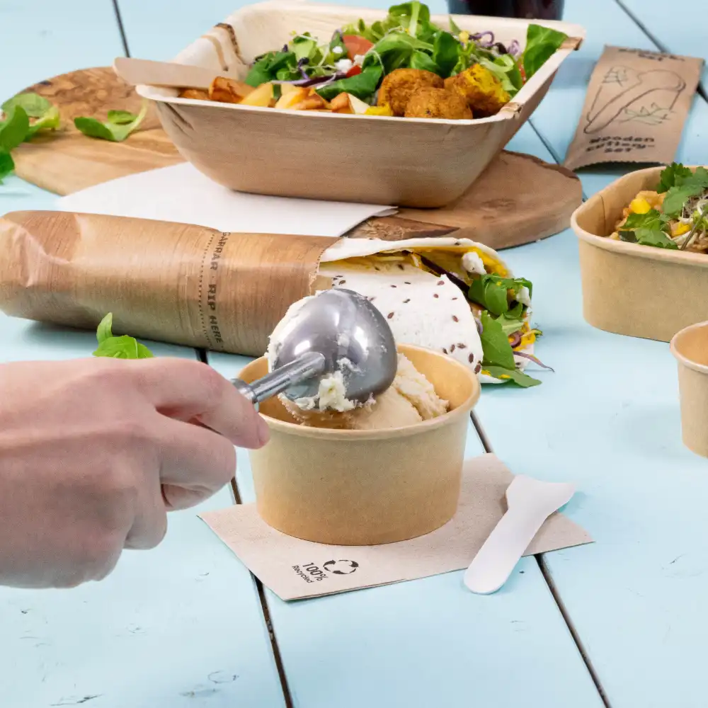 Een hand die een metalen ijslepel gebruikt om uit een bruine papieren beker te serveren; andere voedingsmiddelen en containers staan op de achtergrond op een blauwe tafel.