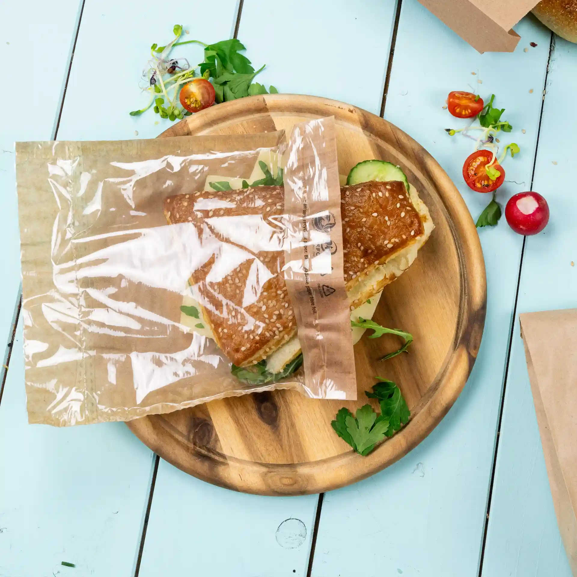 Transparante snackzak met een sandwich erin, geplaatst op een houten bord met verse kruiden en groenten.