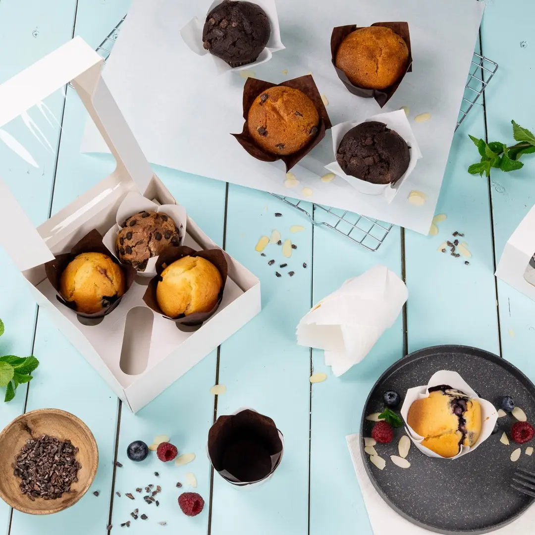 Een witte cupcake-doos met drie muffins, omringd door extra muffins, bessen en chocoladestukjes op een blauwe ondergrond.