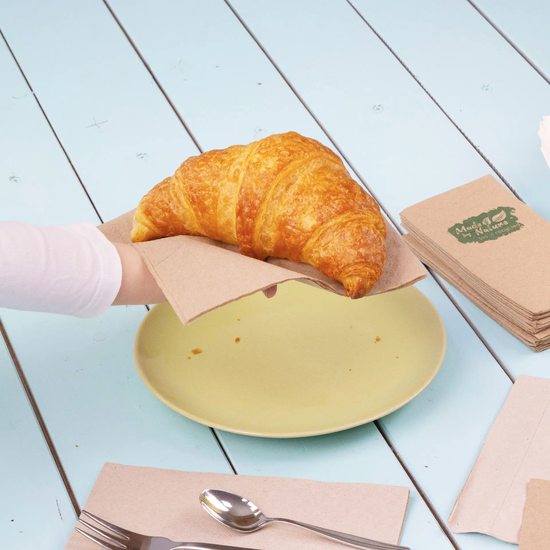 Een hand houdt een croissant op een papieren servet boven een gele plaat, met extra servetten en bestek in de buurt.