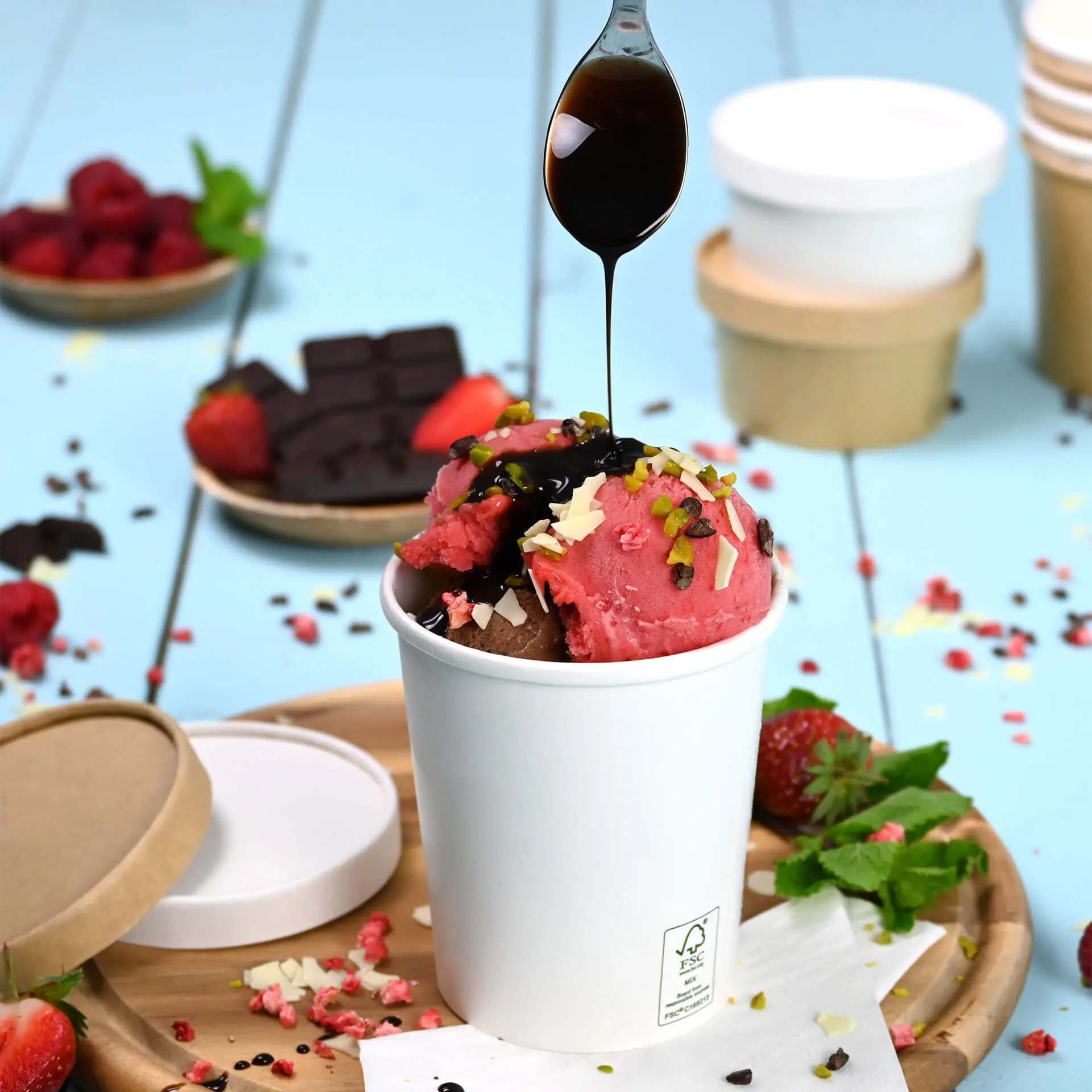 Een witte papieren ijsbekertje gevuld met chocolade- en aardbeienijs, getopt met verschillende toppings en chocoladesaus.