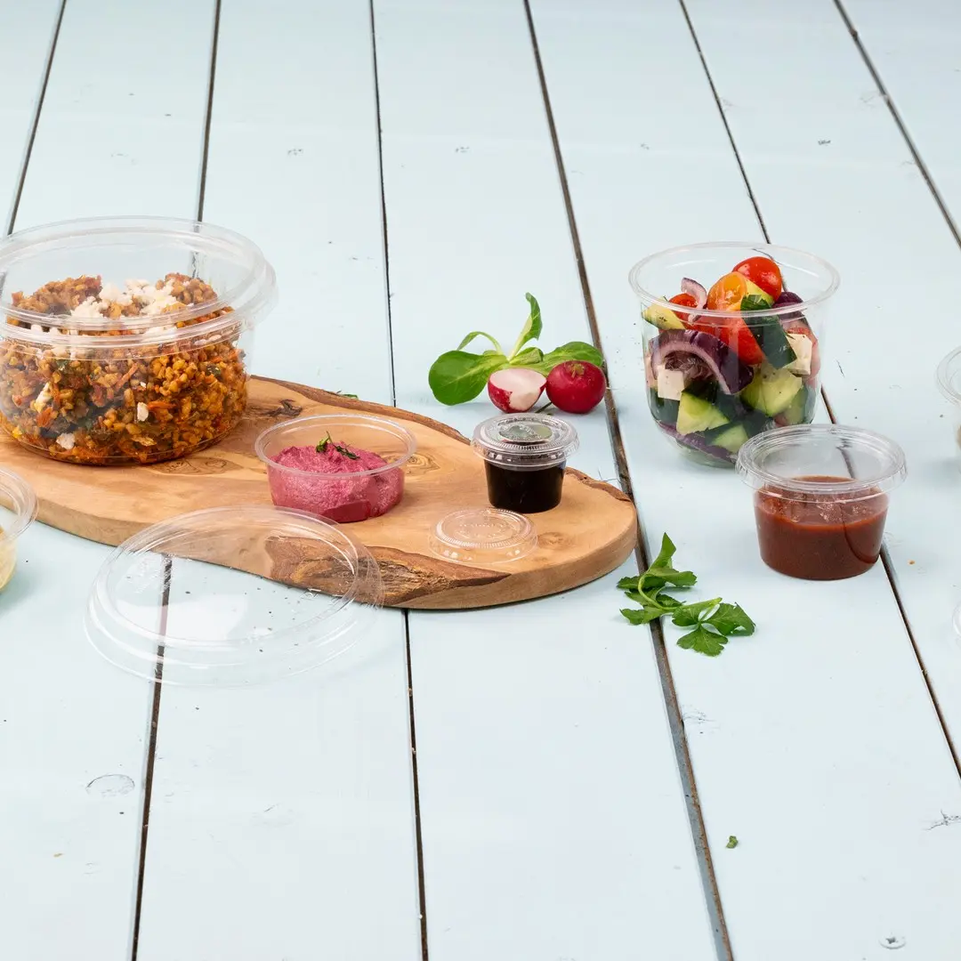 Heldere plastic containers met verschillende voedingsmiddelen, waaronder een salade, dips en een graan gerecht, op een houten plank gerangschikt.