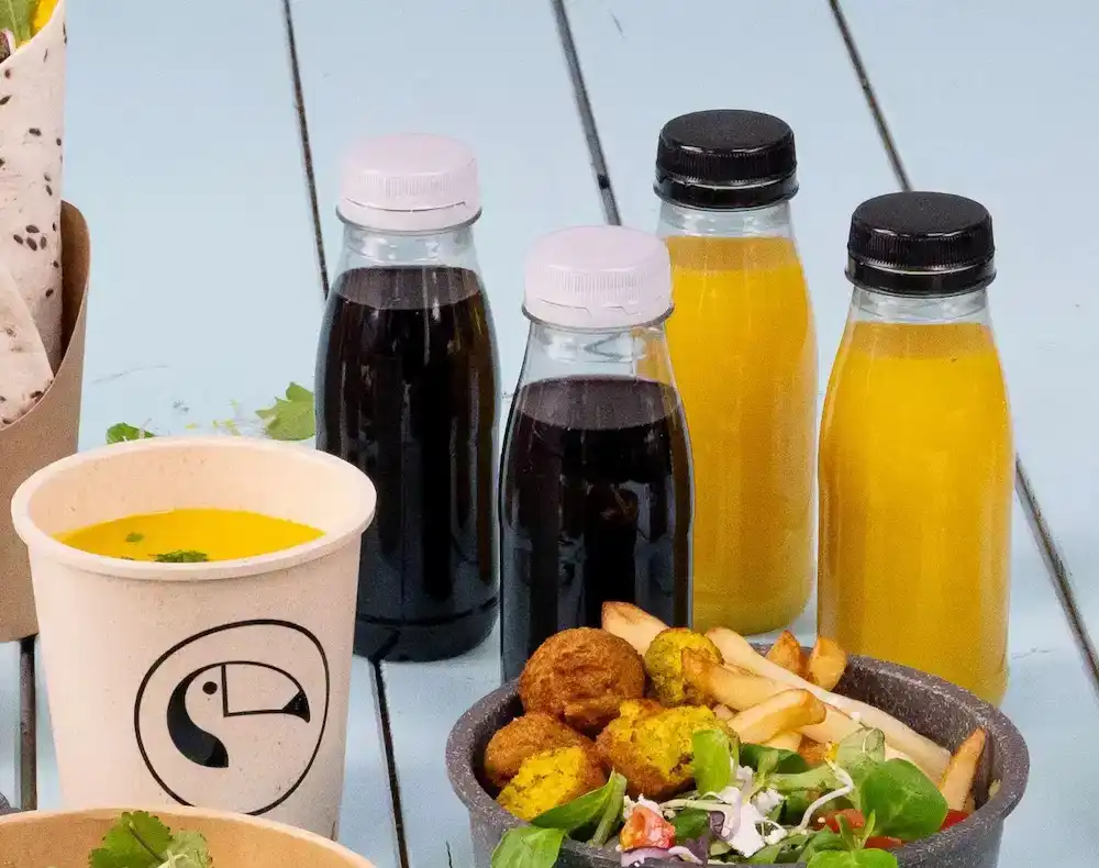 Diverse voedingsmiddelen, waaronder rPET-plastic flessen met verschillende dranken, een beker soep en een kom salade, friet en falafel.