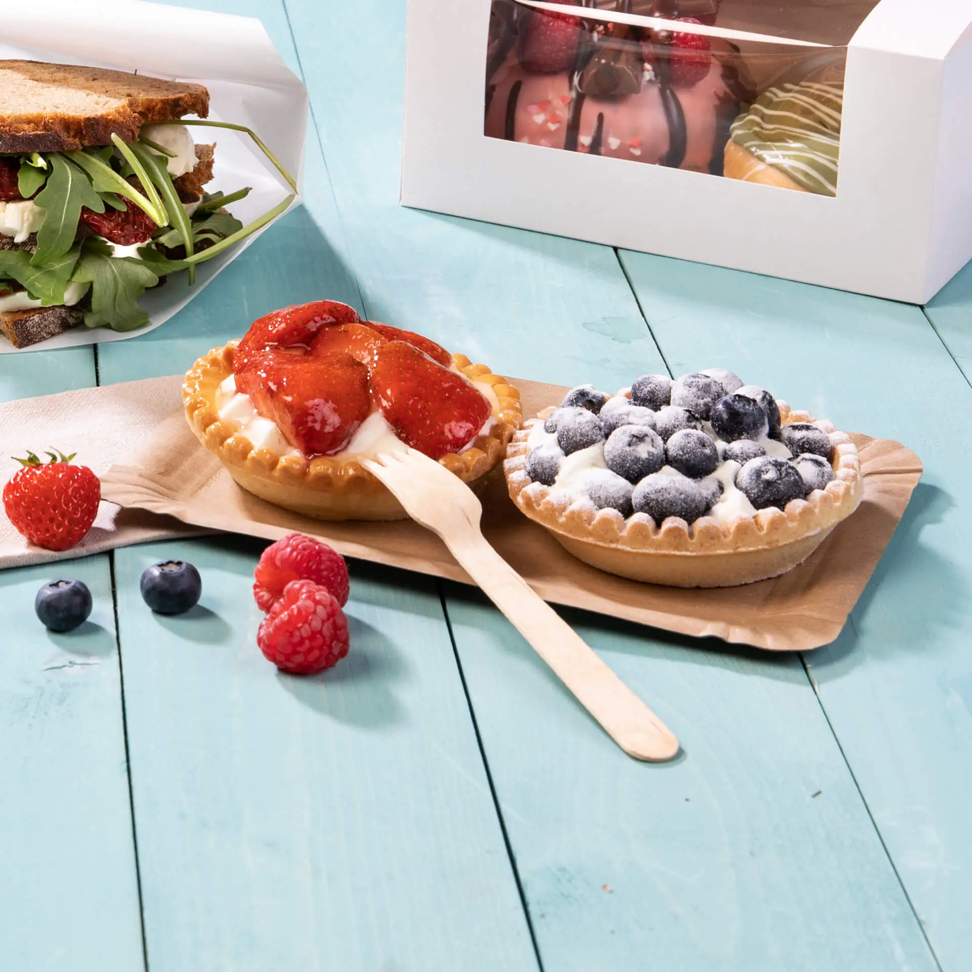 Twee desserttaarten met aardbeien en bosbessen op een bruine kartonnen plaat, naast verse bessen en een sandwich.