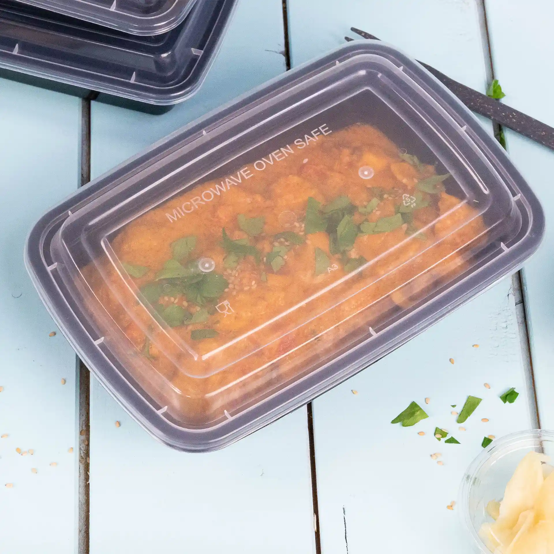 Rechthoekige transparante container met een herbruikbaar deksel, gevuld met voedsel en gegarneerd met groene kruiden, op een blauwe ondergrond.