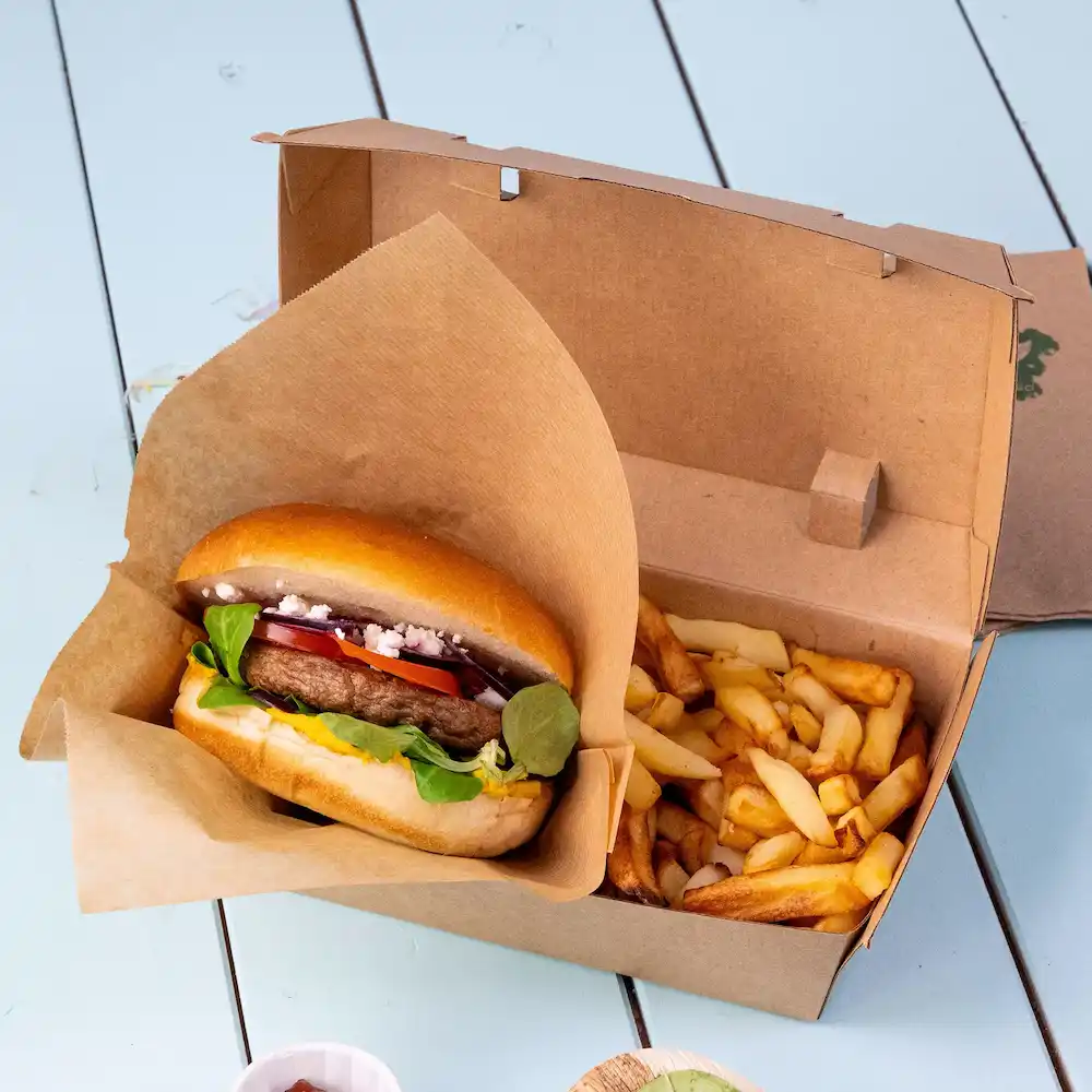 Burger in papierverpakking met verse groenten naast een doos friet op een blauwe ondergrond.