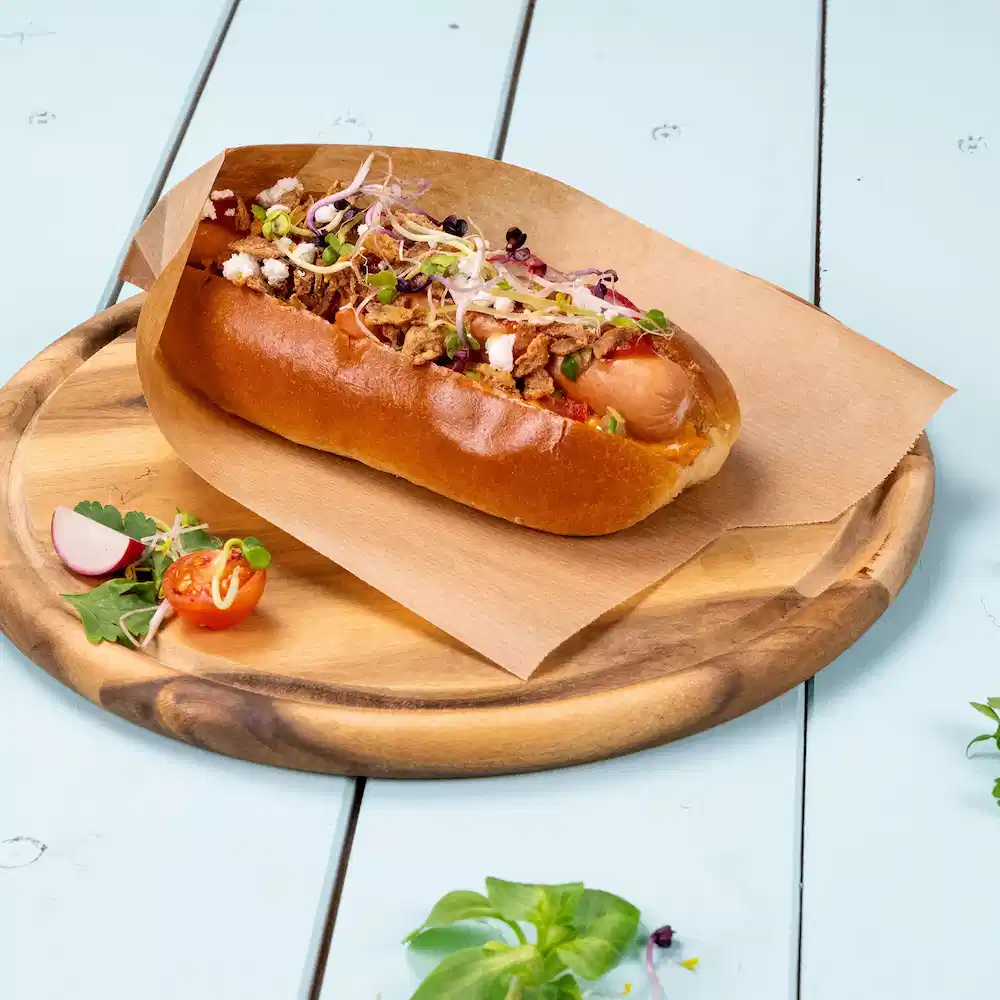 Hotdog in een zacht broodje, belegd met knapperige uien, spruiten en gegarneerd met cherrytomaten en radijs op een houten bord.