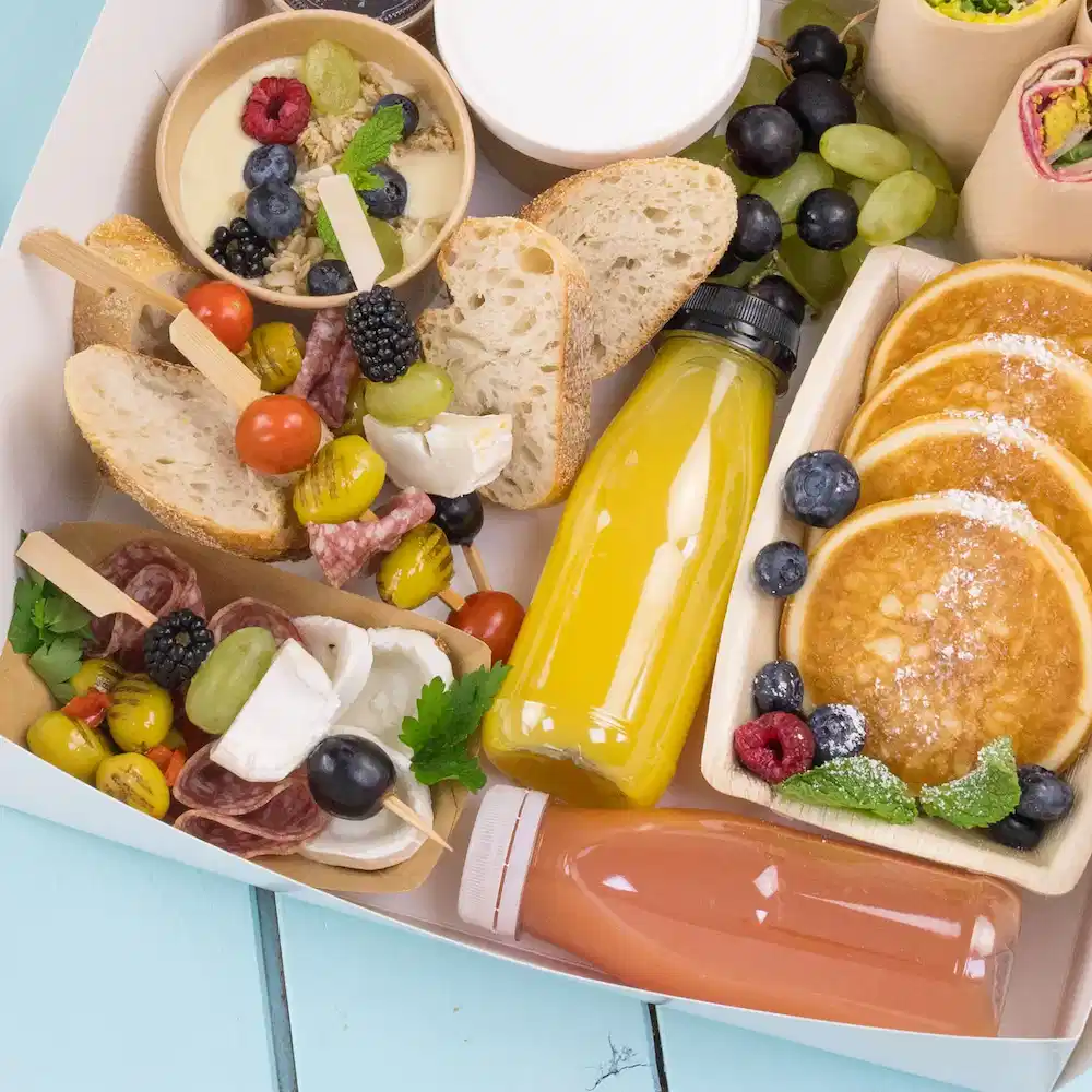 Een verscheidenheid aan voedingsmiddelen, waaronder pannenkoeken, fruit, brood en dranken, in een container op een lichtblauwe ondergrond.