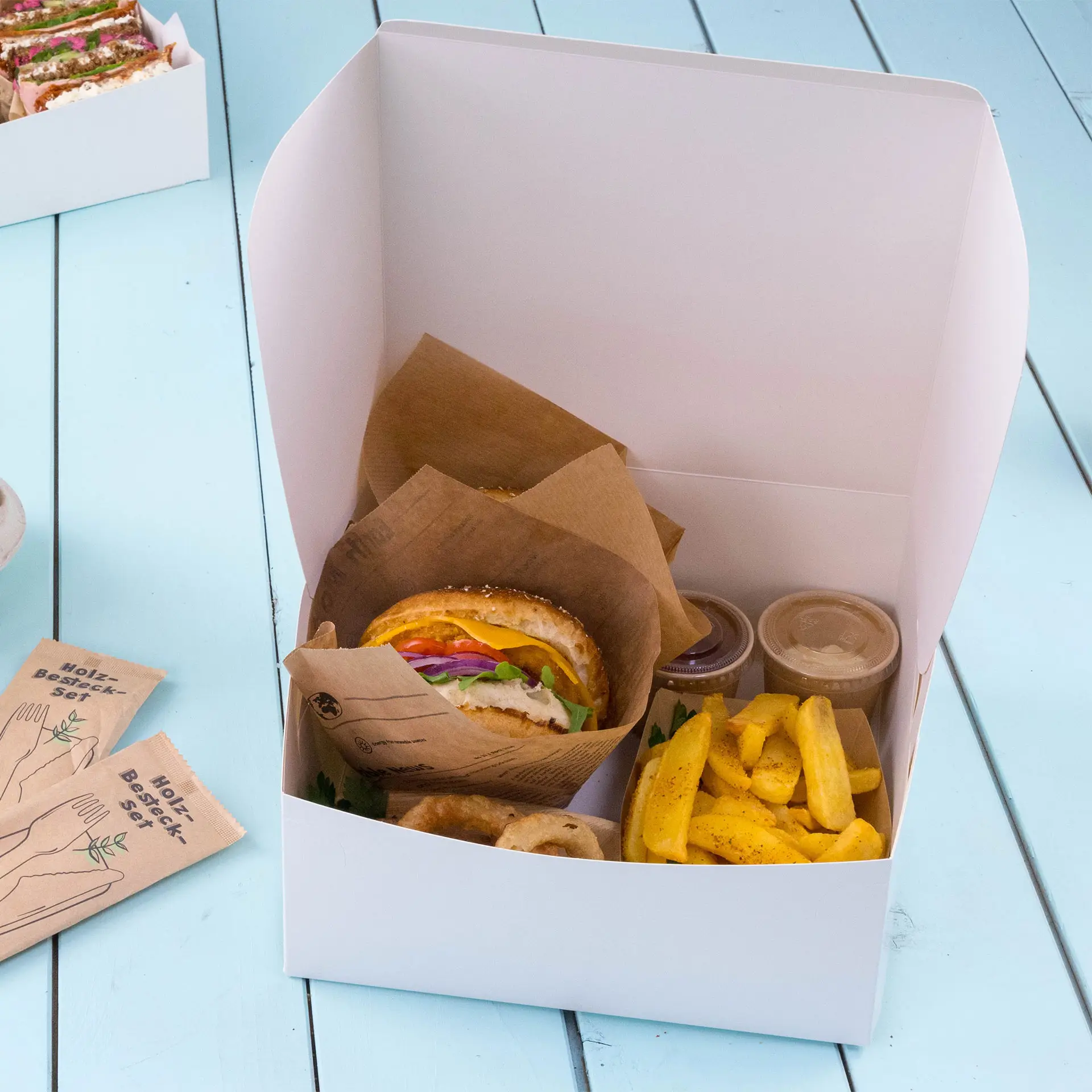 Witte kartonnen taartdoos met een sandwich, friet en twee sauscontainers, met een gevouwen deksel.