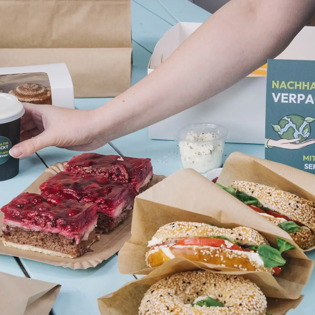 Een hand reikt naar een beker naast een bord met gelaagd dessert, twee sesam-bagels met vullingen en een container room.