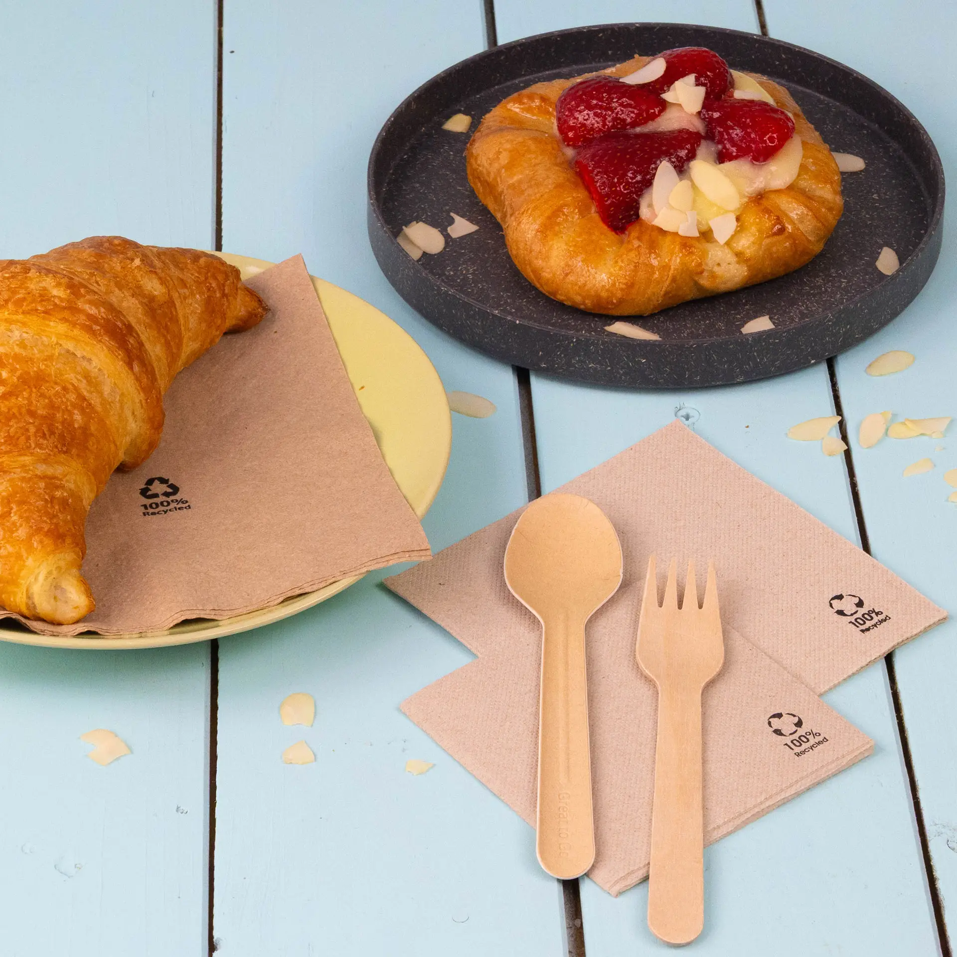 Een witte koffiekop met een koekje op een wit bord, omringd door een kommetje frambozen en cocktailservetten van gerecycled papier.