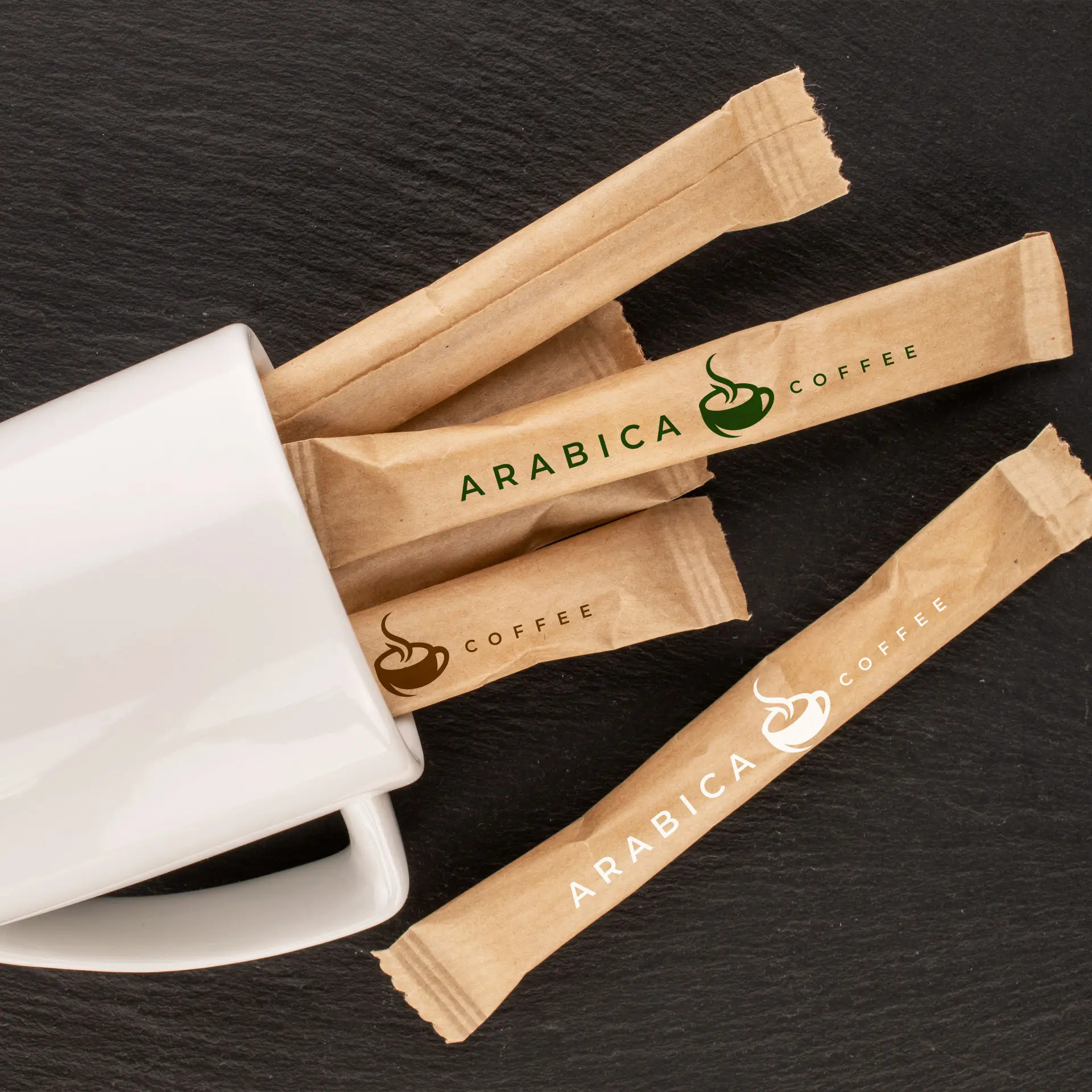 Bruine biologische rietsuikersticks in kraftpapier met het "ARABICA COFFEE"-logo, naast een witte mok op een donkere ondergrond.