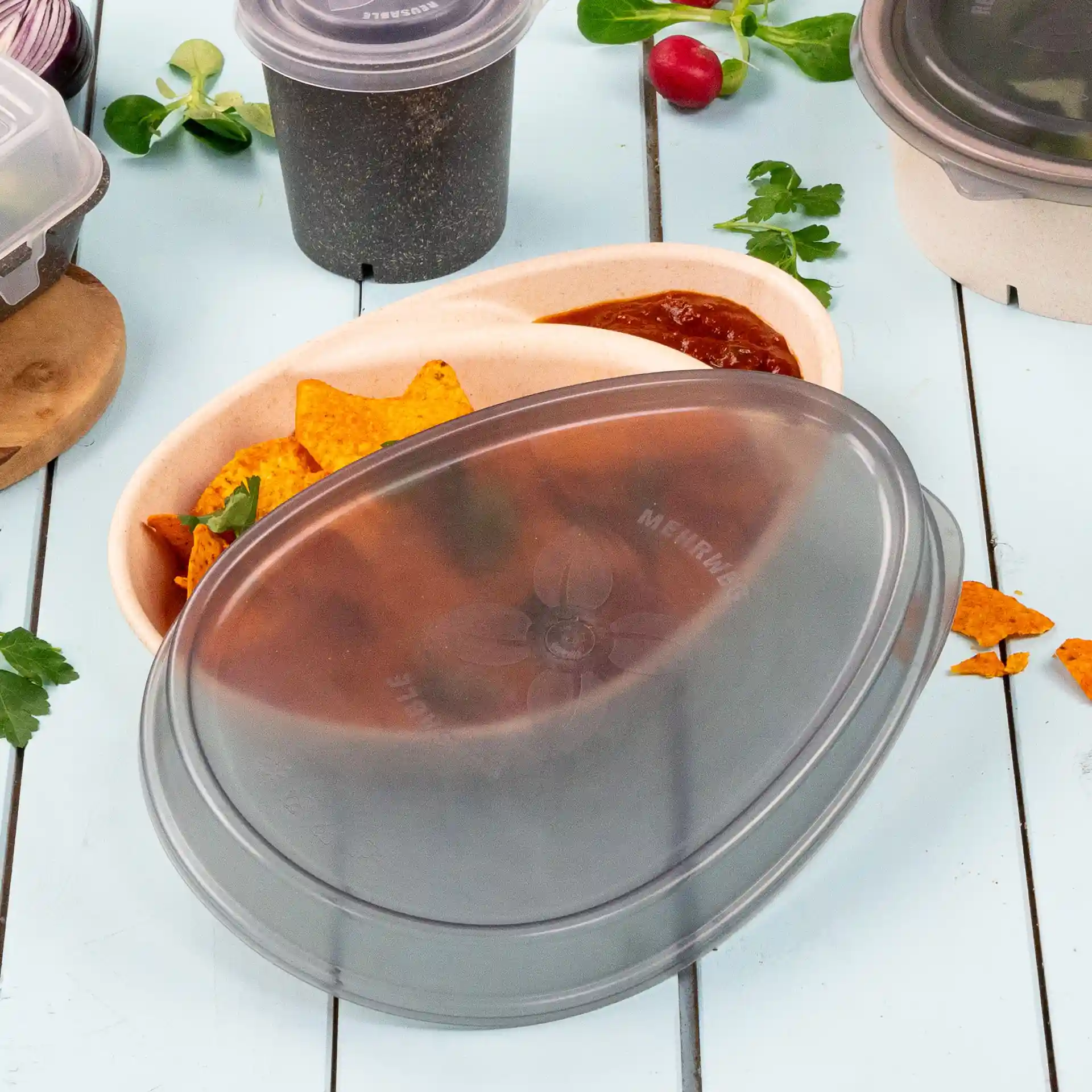 Grijze ovale herbruikbare deksel voor een container, geplaatst op een kom met nacho's en salsa, op een lichtblauwe ondergrond.