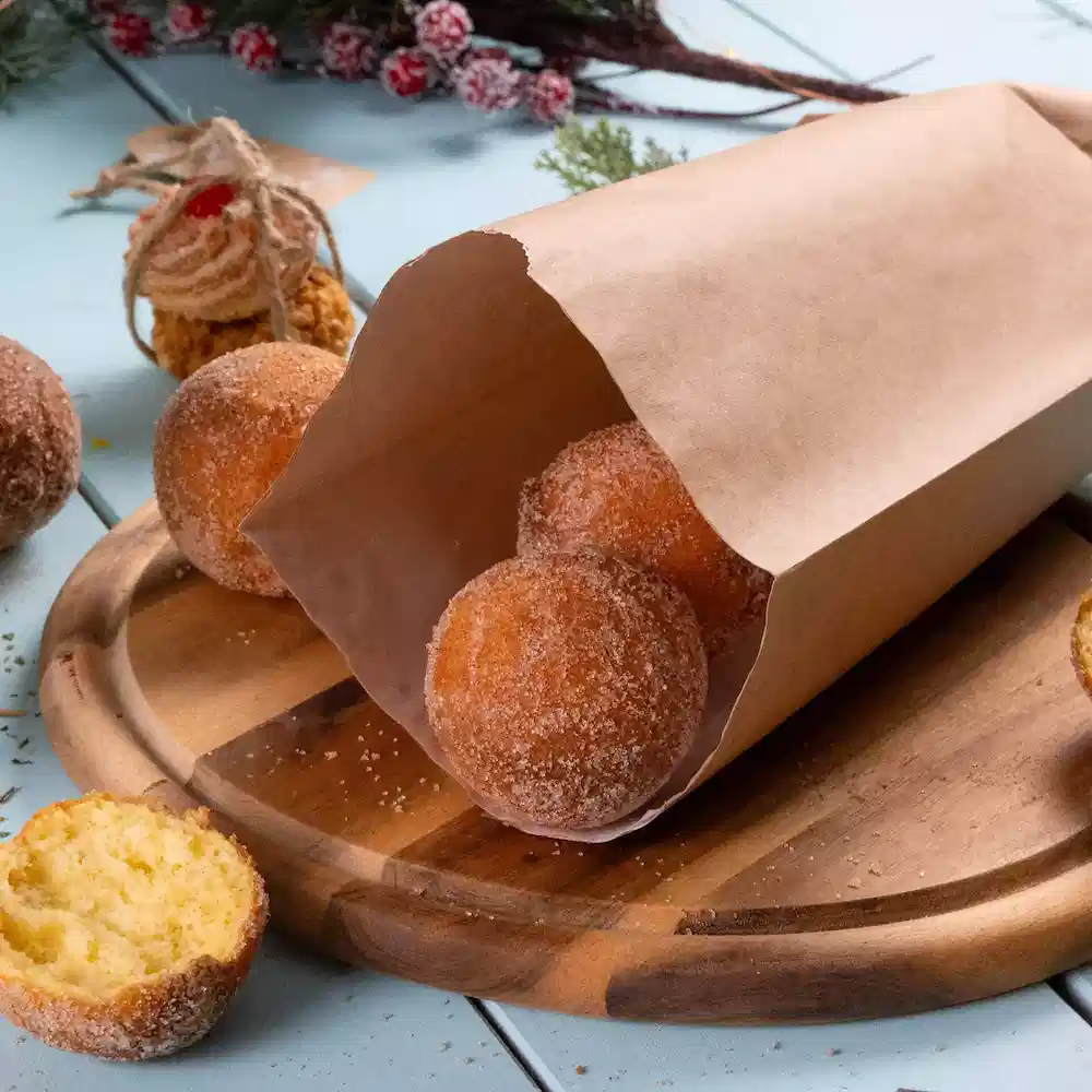 Een bruine papieren tas bevat meerdere ronde donuts met poedersuiker op een houten snijplank, met een donut gedeeltelijk gesneden.
