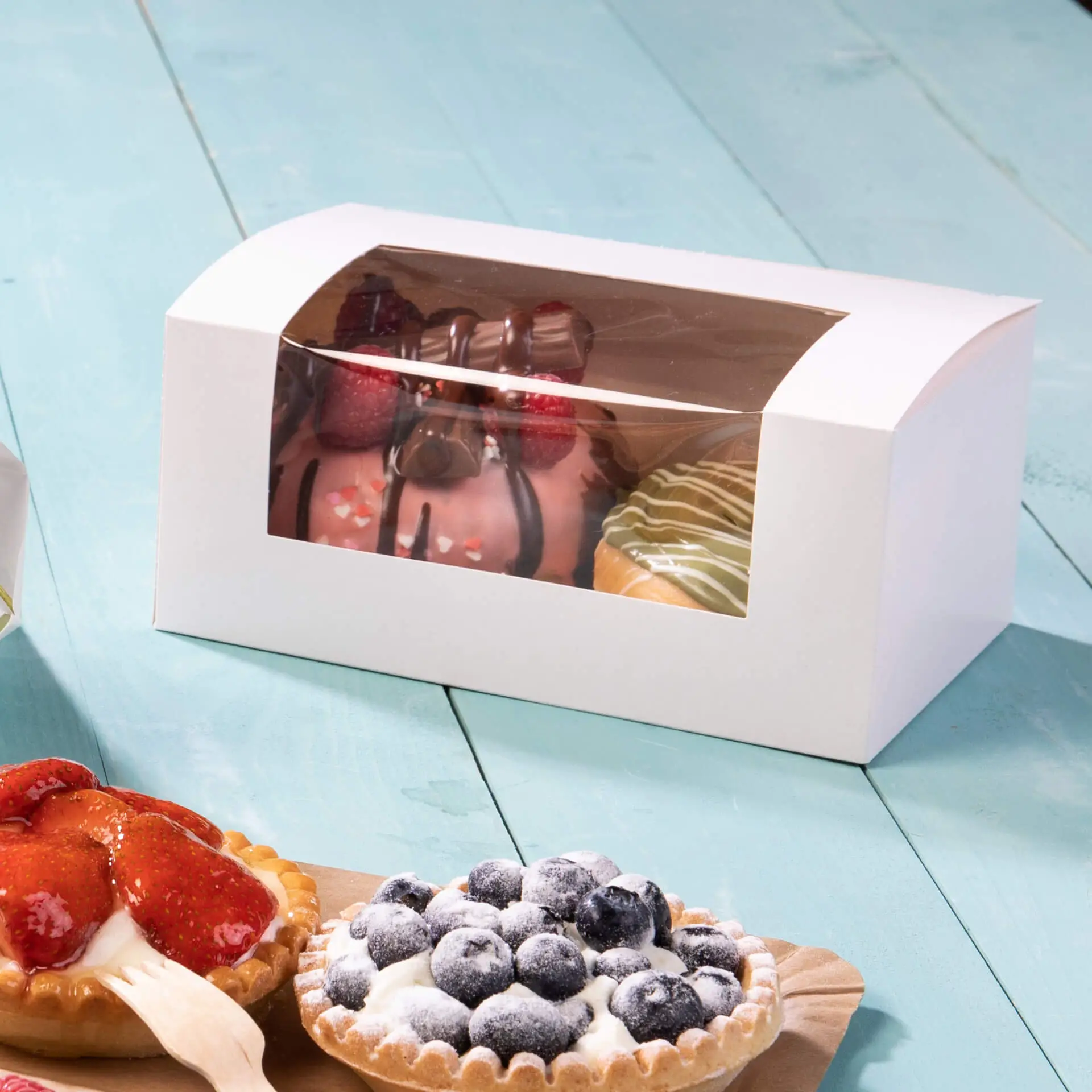 Witte cupcake-box met een cellulosevenster dat diverse gebakjes toont, waaronder taarten en tarts, op een turquoise ondergrond.