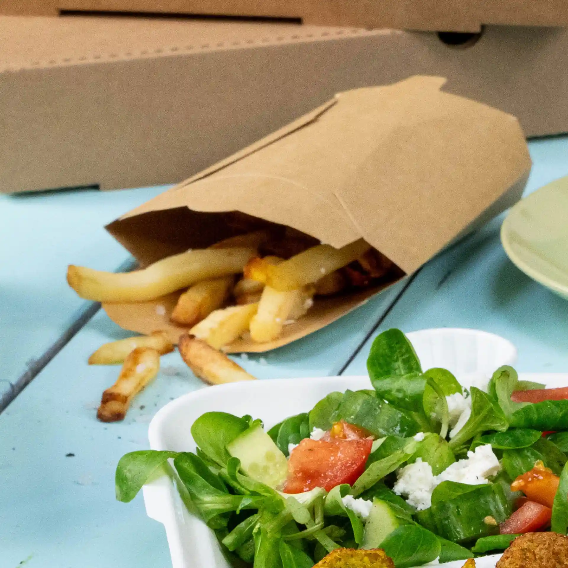 Bruine kartonnen chip-schep met frietjes die eruit vallen, naast een salade in een witte container op een blauwe ondergrond.
