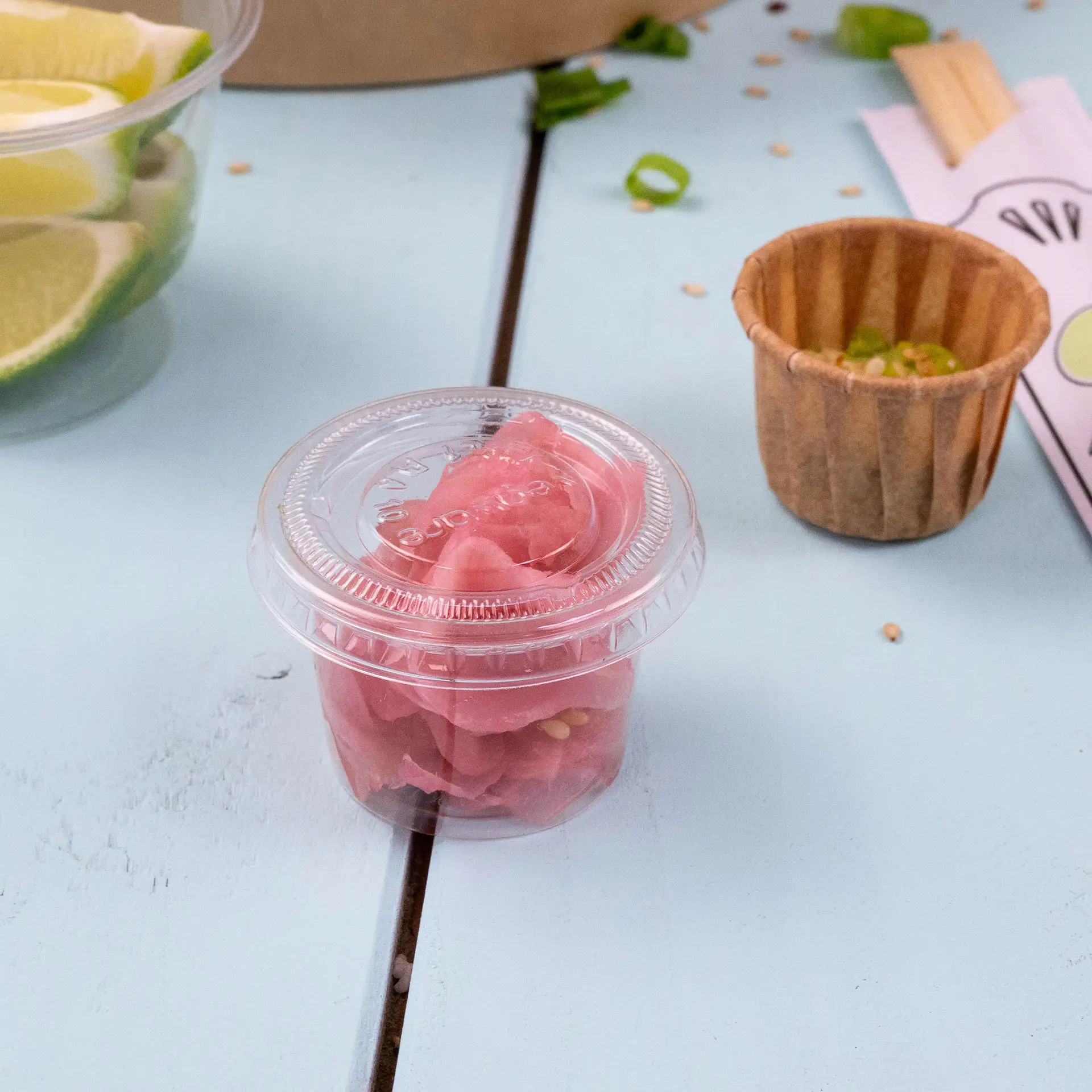 Een heldere plastic beker met roze gemarineerde gember op een lichtblauwe ondergrond, met een bruine papieren beker en limoenschijfjes op de achtergrond.