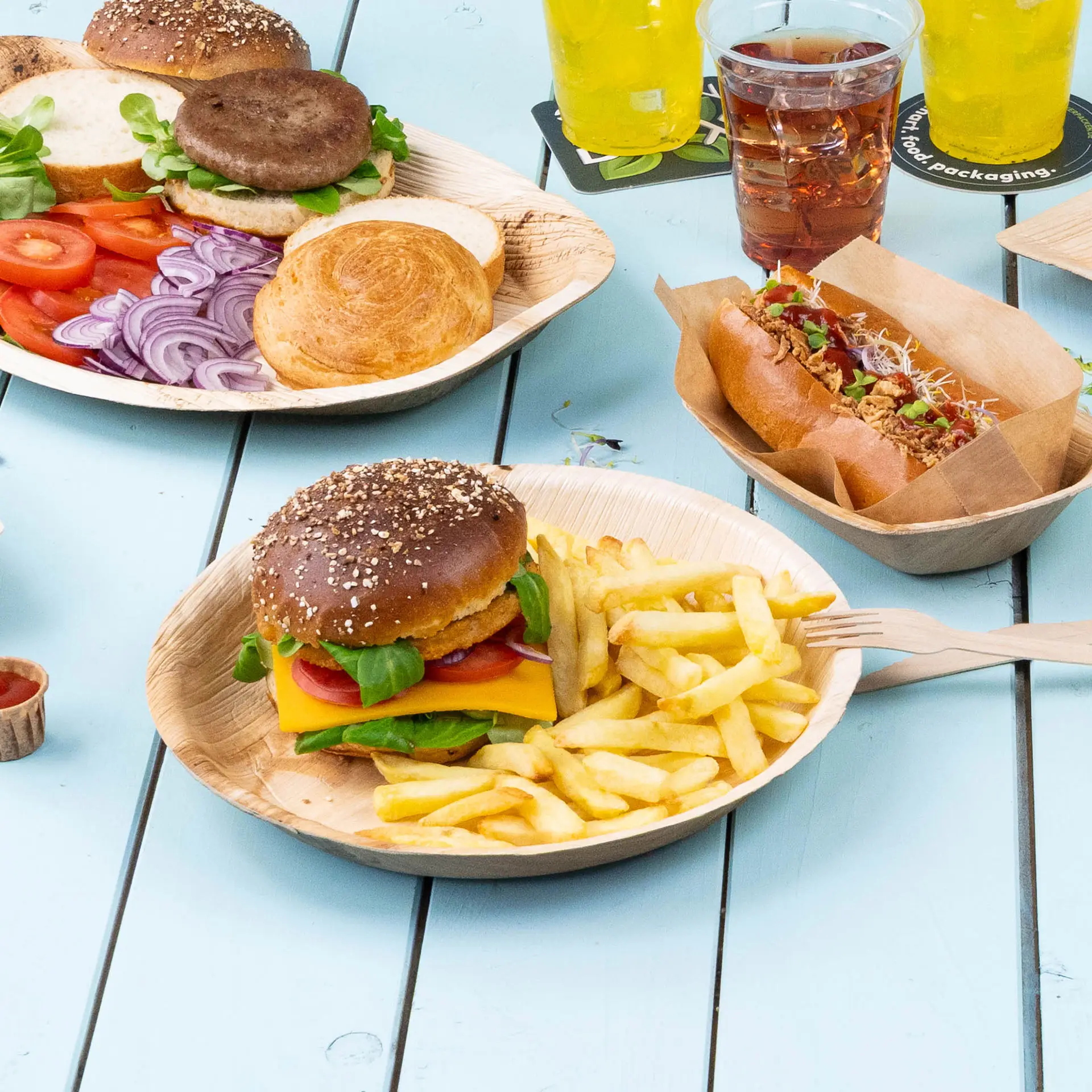 Een palmbladplaat bevat een cheeseburger met sla, tomaat en frietjes, naast plakjes groenten en een hotdog.
