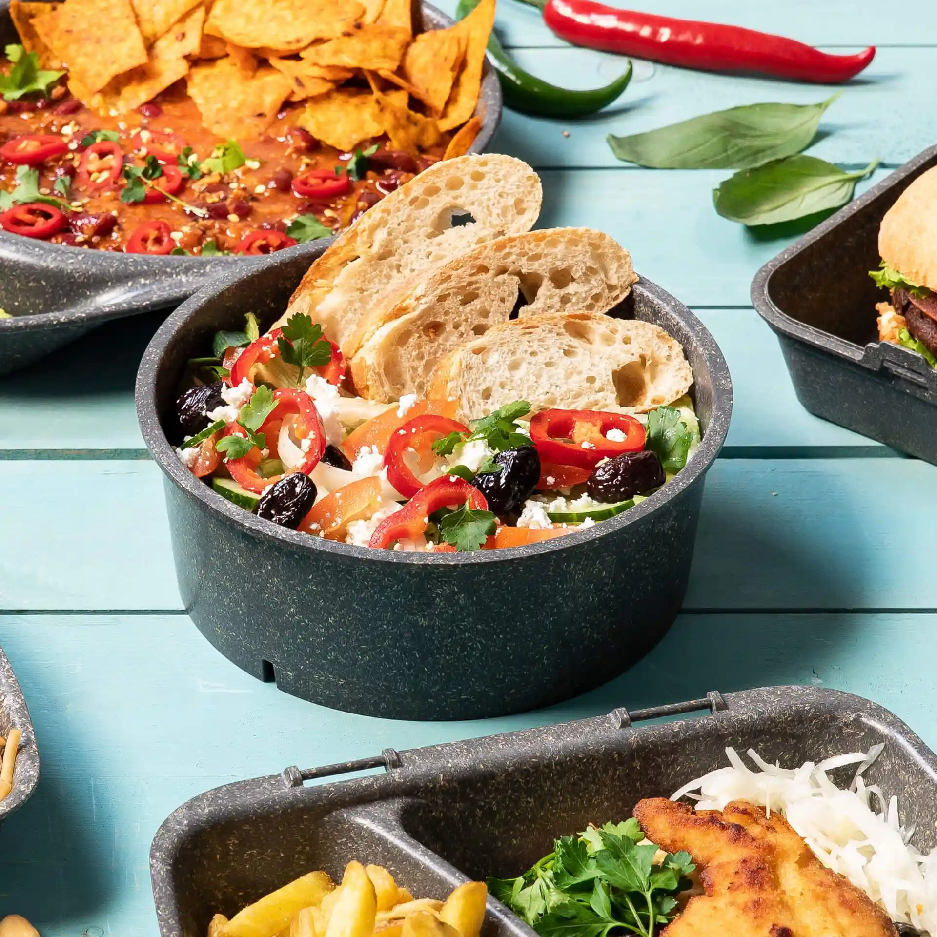 Ronde grijze herbruikbare kom gevuld met salade, getopt met gesneden brood, paprika en kruiden, op een blauwe houten ondergrond.