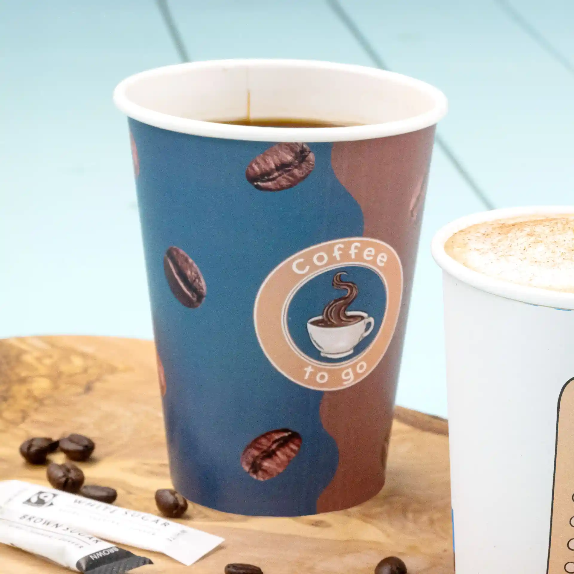 Papieren beker met gedrukt ontwerp, gevuld met koffie, op een houten oppervlak met suikerzakjes en koffiebonen.