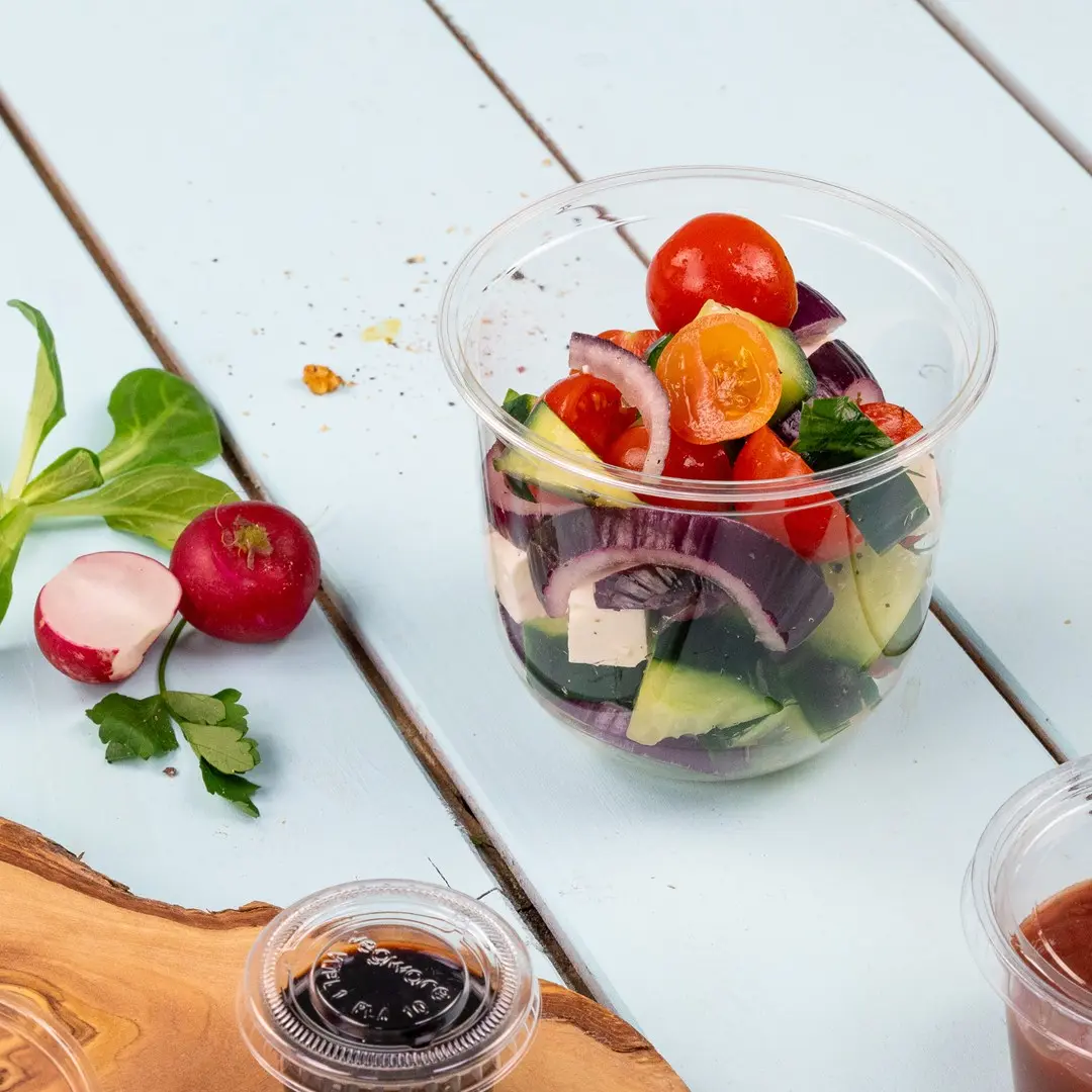 Heldere plastic beker gevuld met gemengde salade met cherrytomaten, komkommer, rode ui en feta, met radijzen erbij.