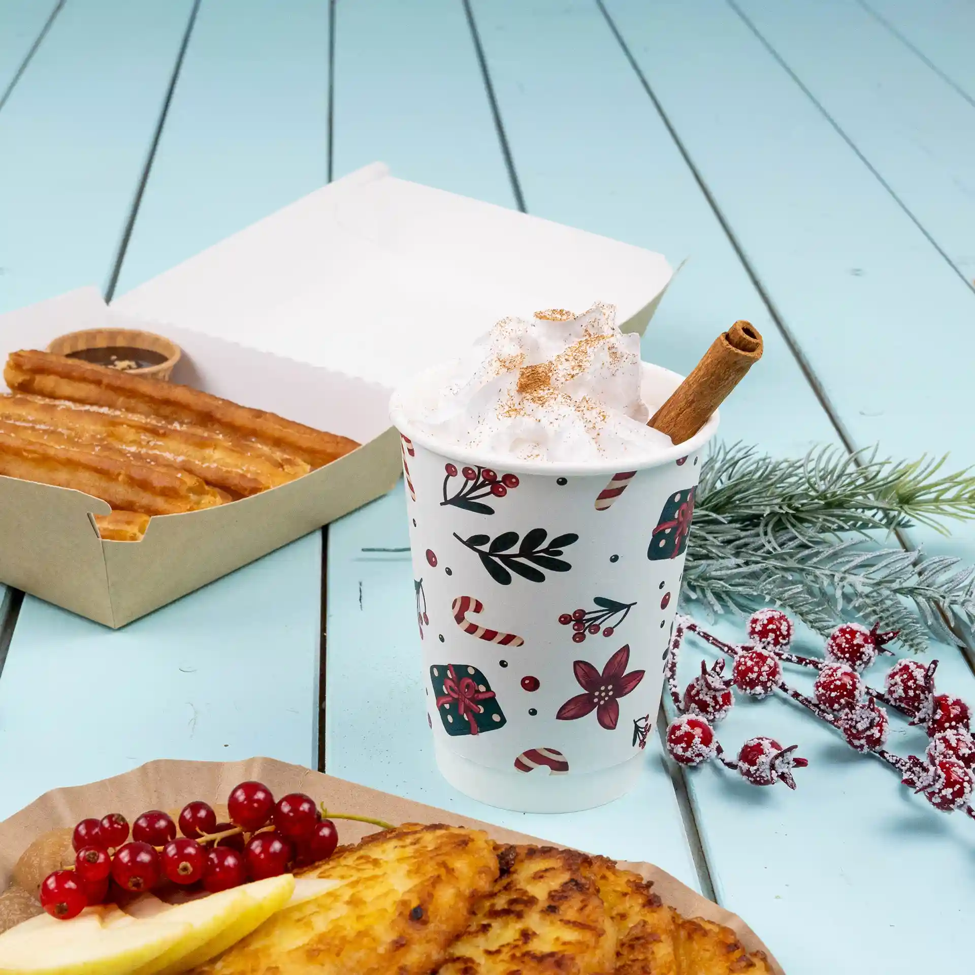Dubbelwandige beker met winterontwerp, gevuld met slagroom en een kaneelstokje, naast churros en fruit.