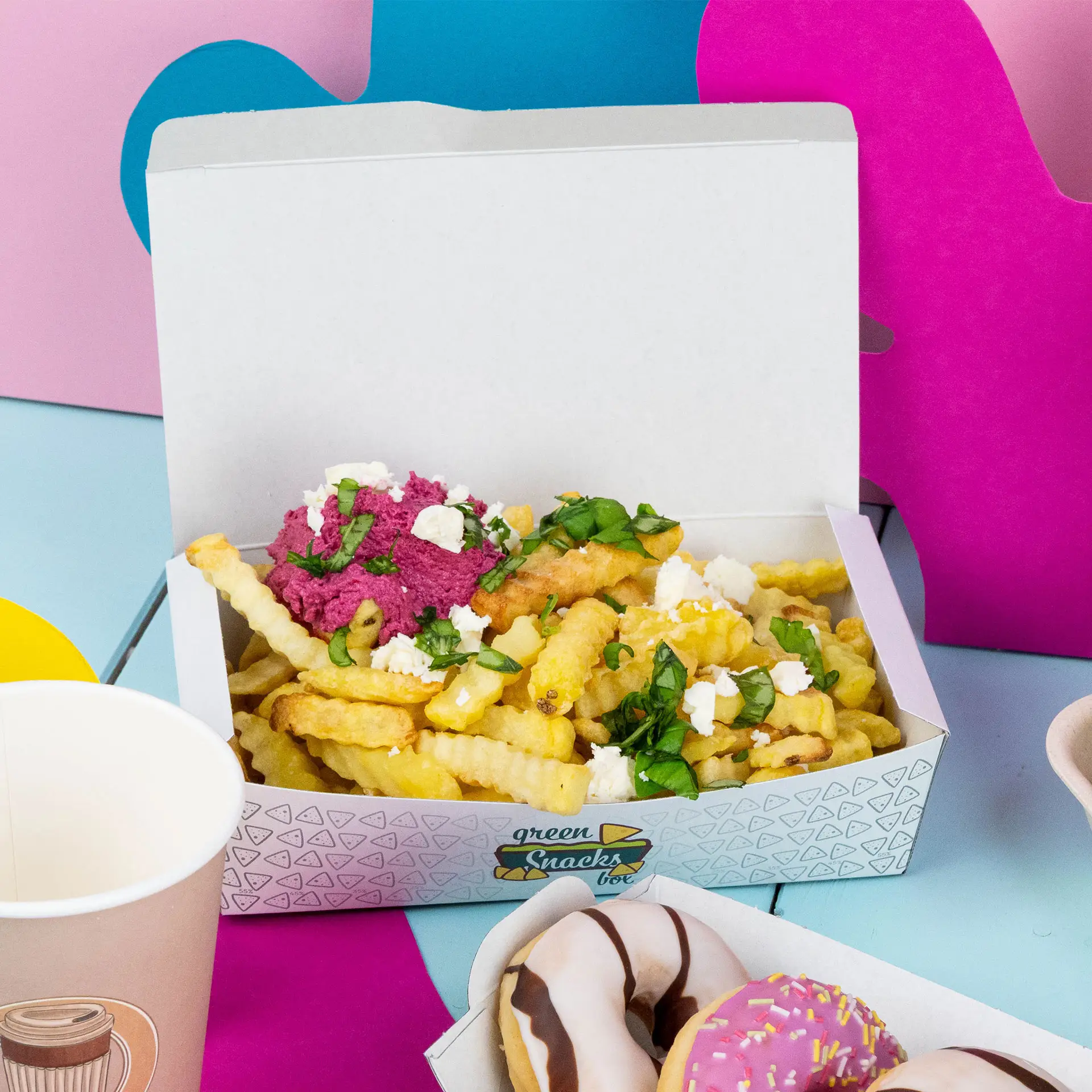Witte snackdoos met gekrulde frietjes, roze dip, kruiden en kaas, met donuts en een beker in de buurt.