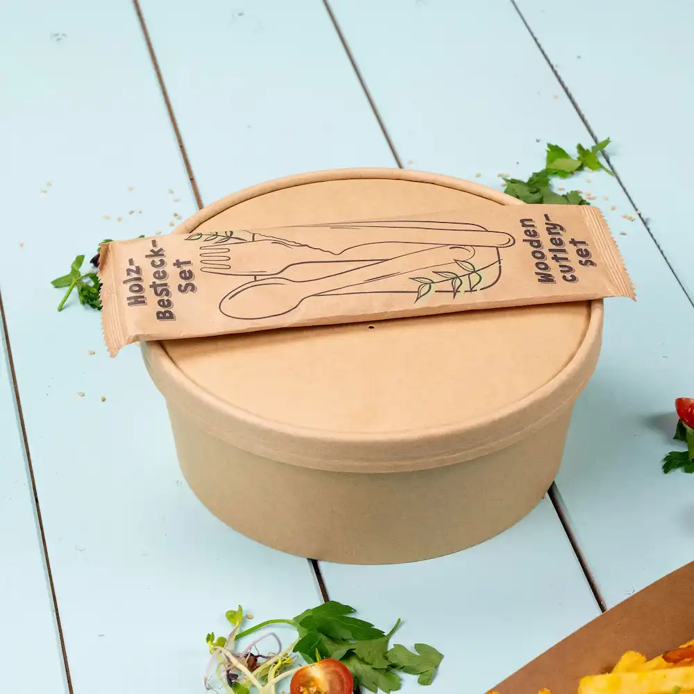 Bruine container met een deksel en een verpakt houten bestekset bovenop, met groene kruiden en salade-elementen in de buurt.