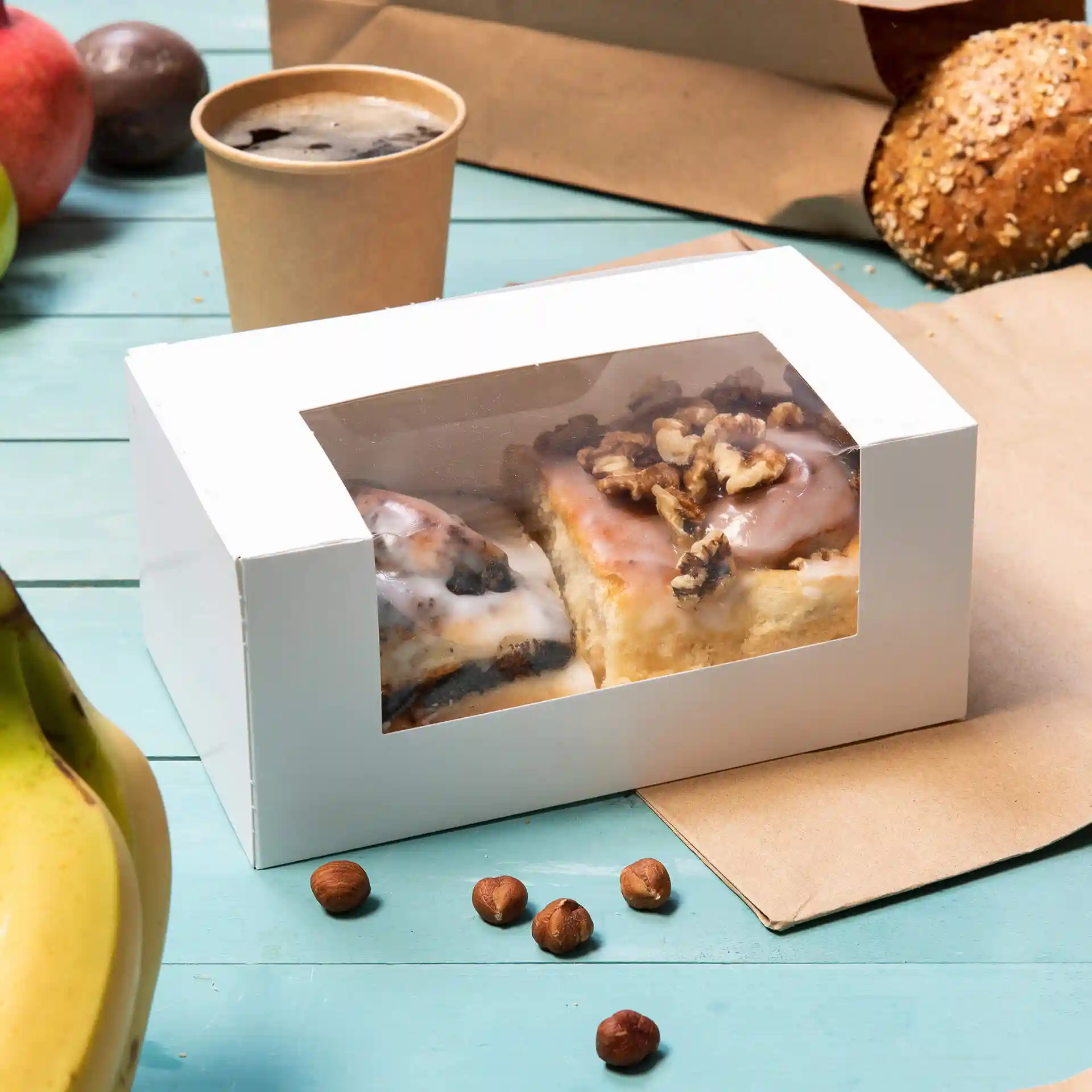 Witte gebakdoos met een cellulosevenster met twee gebakjes, omringd door een banaan, hazelnoten en een beker.