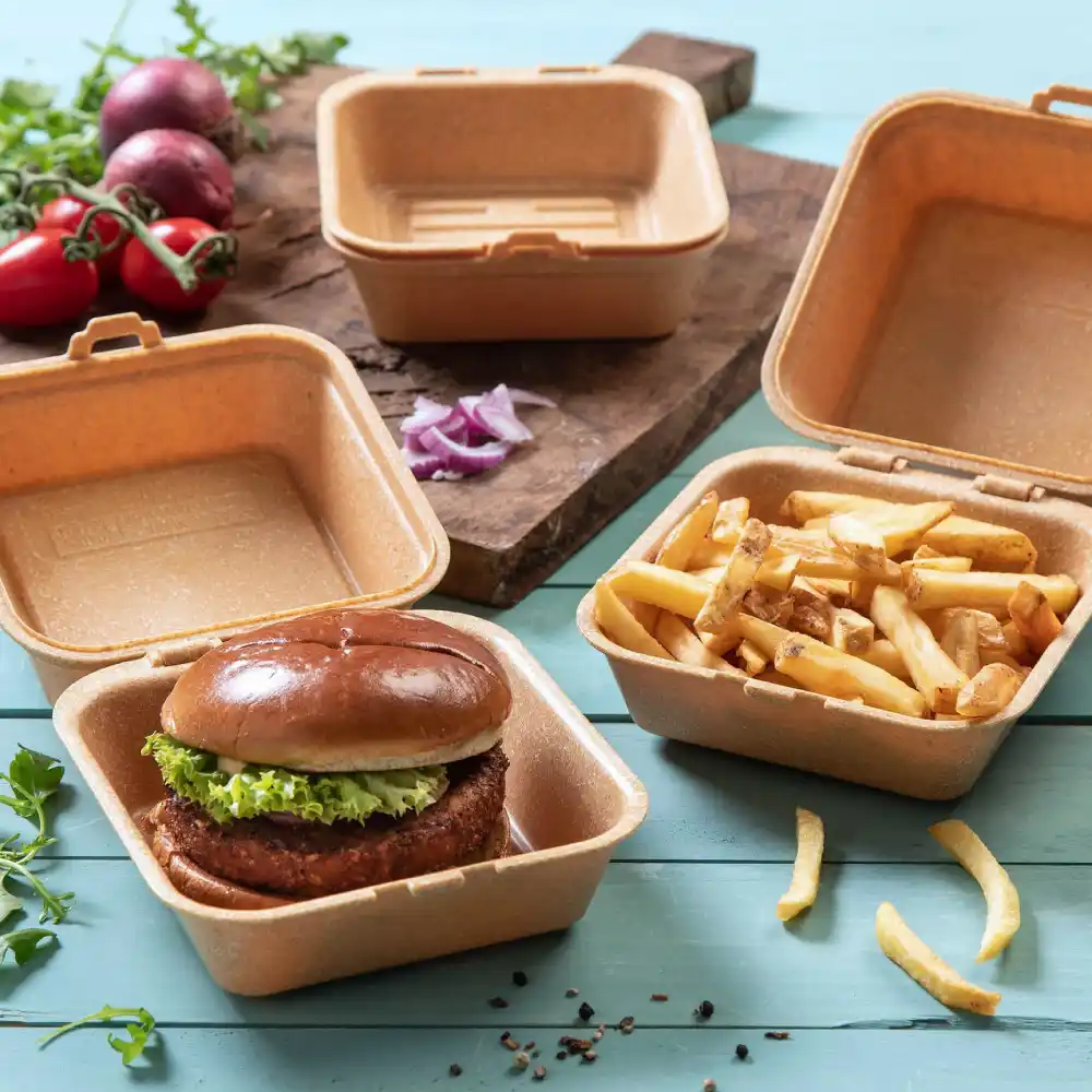 Container met een burger belegd met sla, naast een container met friet, op een houten oppervlak met groenten in de buurt.