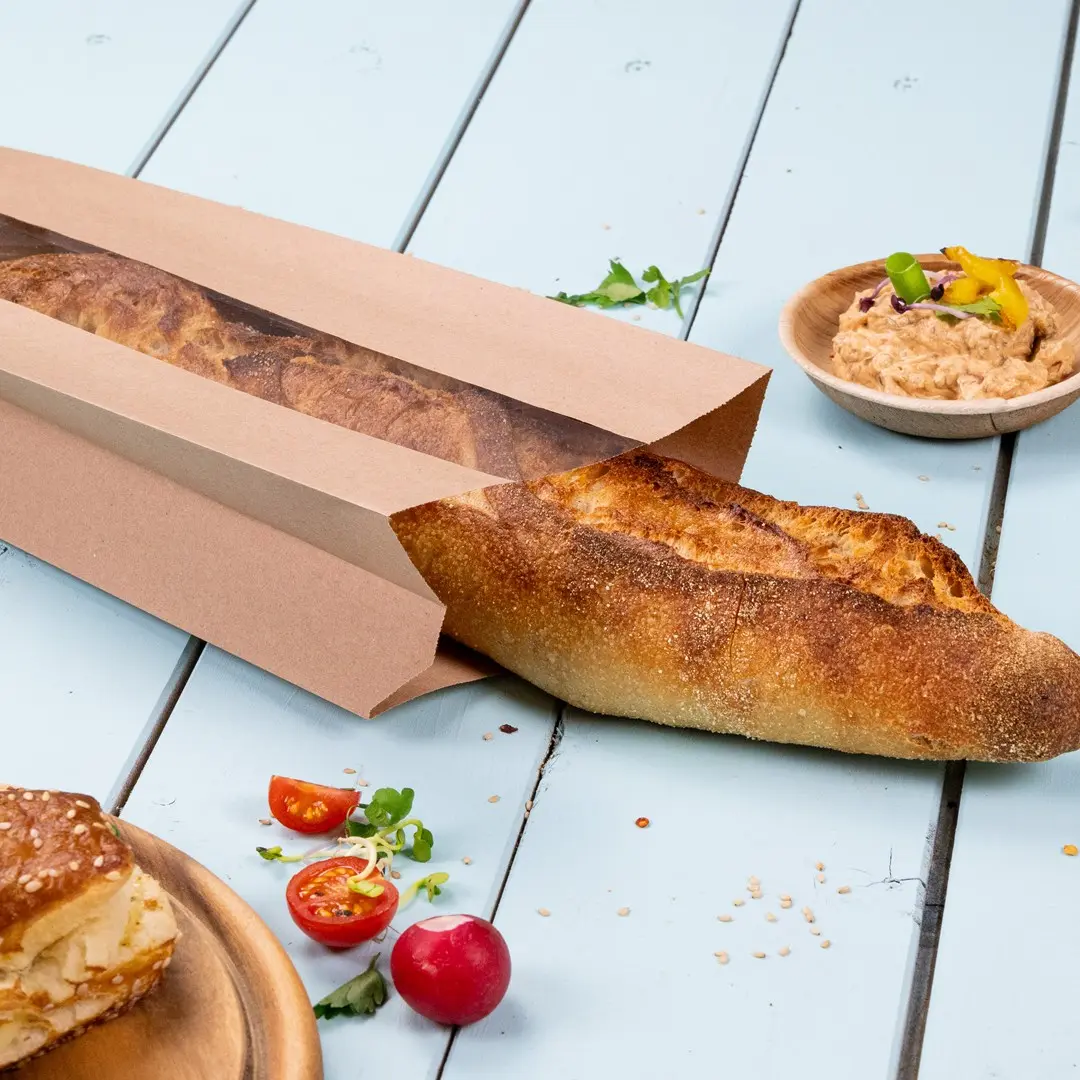 Bruine papieren tas gedeeltelijk open met een baguette, cherrytomaten, radijs en een kom dip op een lichtblauwe ondergrond.