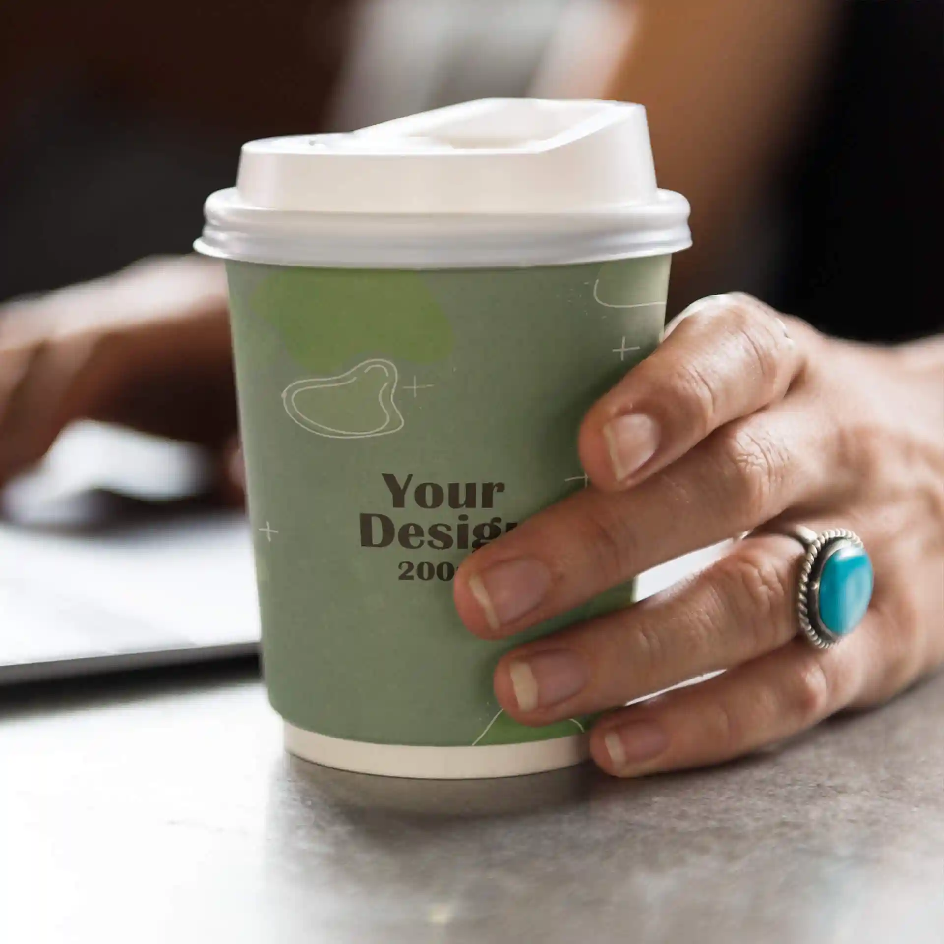 Hand houdt een dubbelwandige papieren beker vast met een groen ontwerp, een witte deksel en een ring om de vinger.