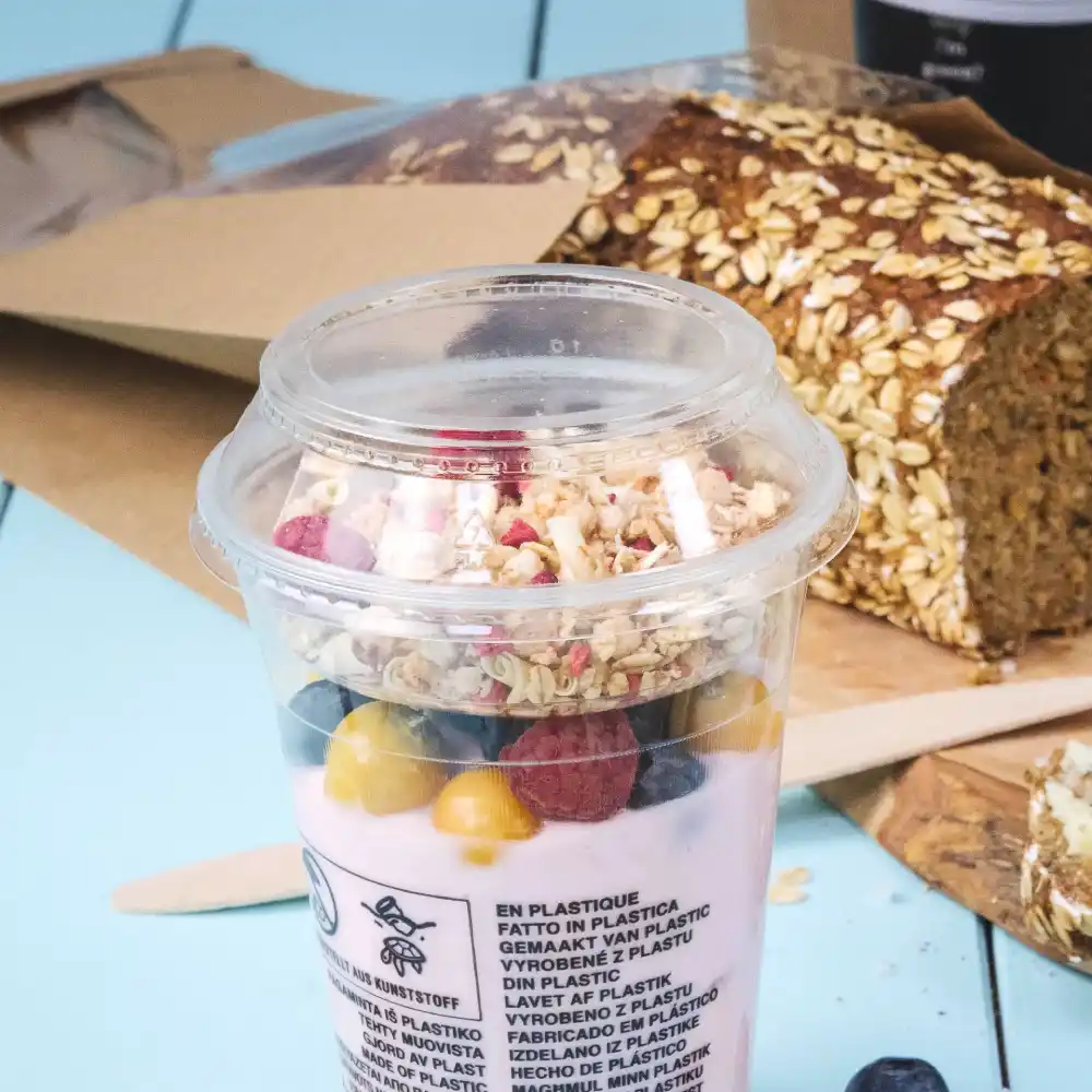 Beker met yoghurt, granola en bessen, naast een brood van haver op een blauwe ondergrond.